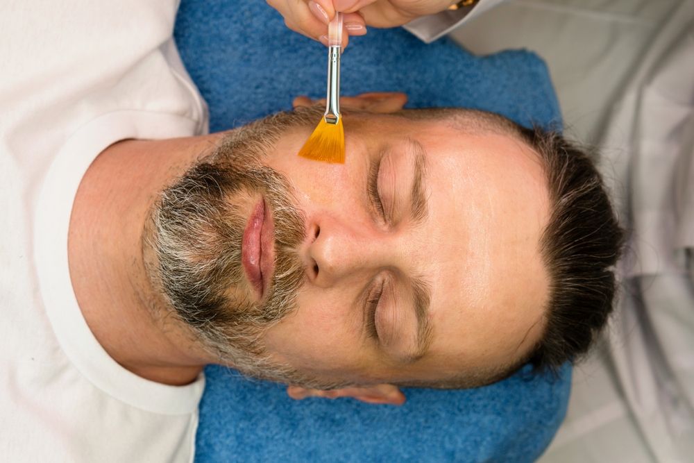 A person lying down receiving a facial treatment, with a small yellow brush applying product to their cheek.