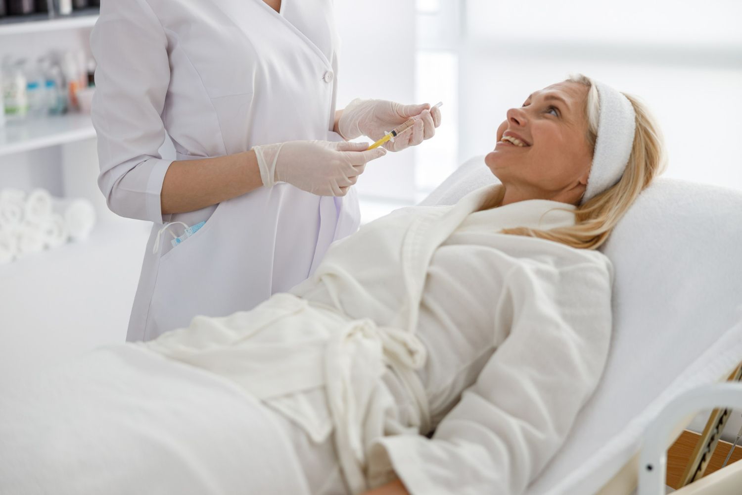 A practitioner wearing gloves prepares a cosmetic injection for a patient resting in a robe on a clinic treatment table.