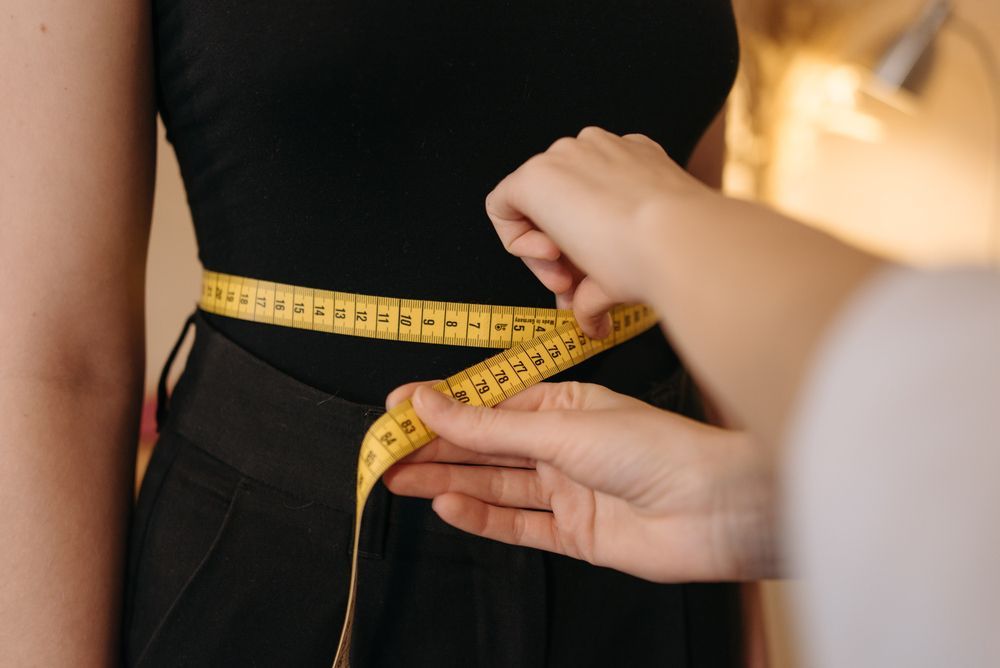 Hands measure a person's waistline with a yellow tape measure over black clothing.