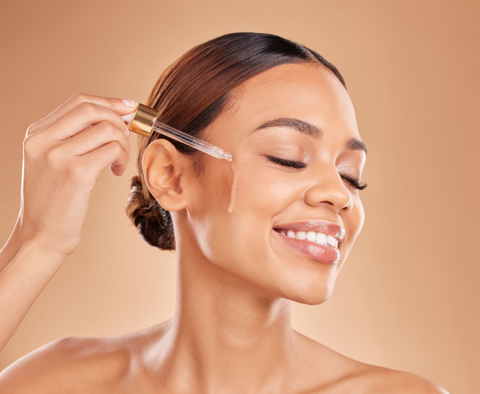 Woman applying product with dropper to her face, smiling with eyes closed against a neutral backdrop.
