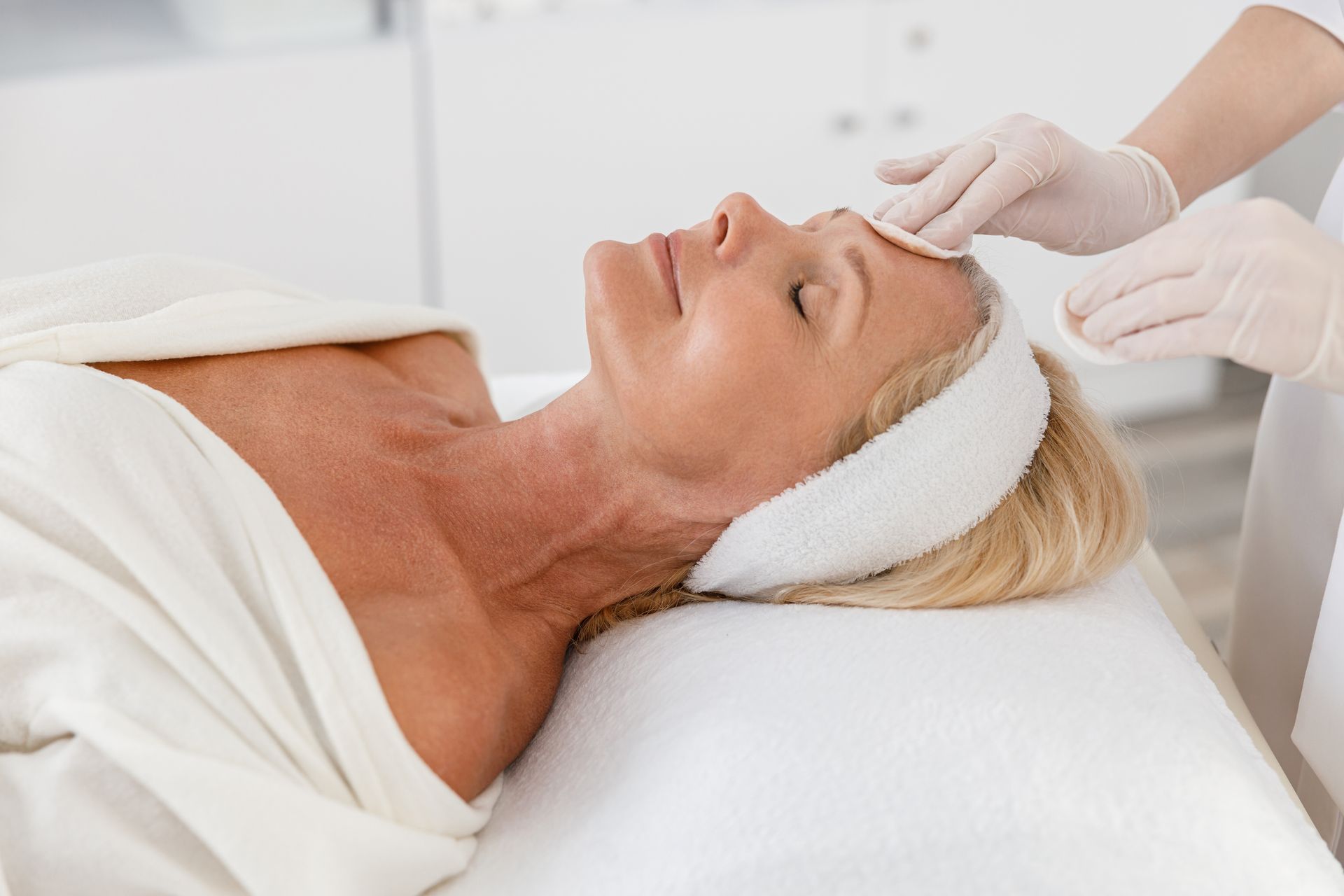 Woman receiving facial treatment in a spa, lying on a white bed. A professional in gloves cleanses her face with cotton pads.