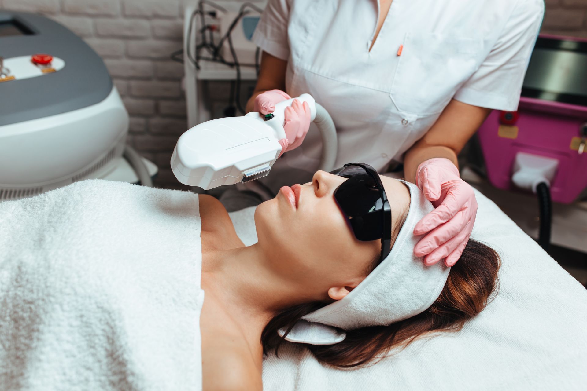 Woman undergoing facial laser treatment in a medical spa setting, wearing protective glasses and a towel head wrap.