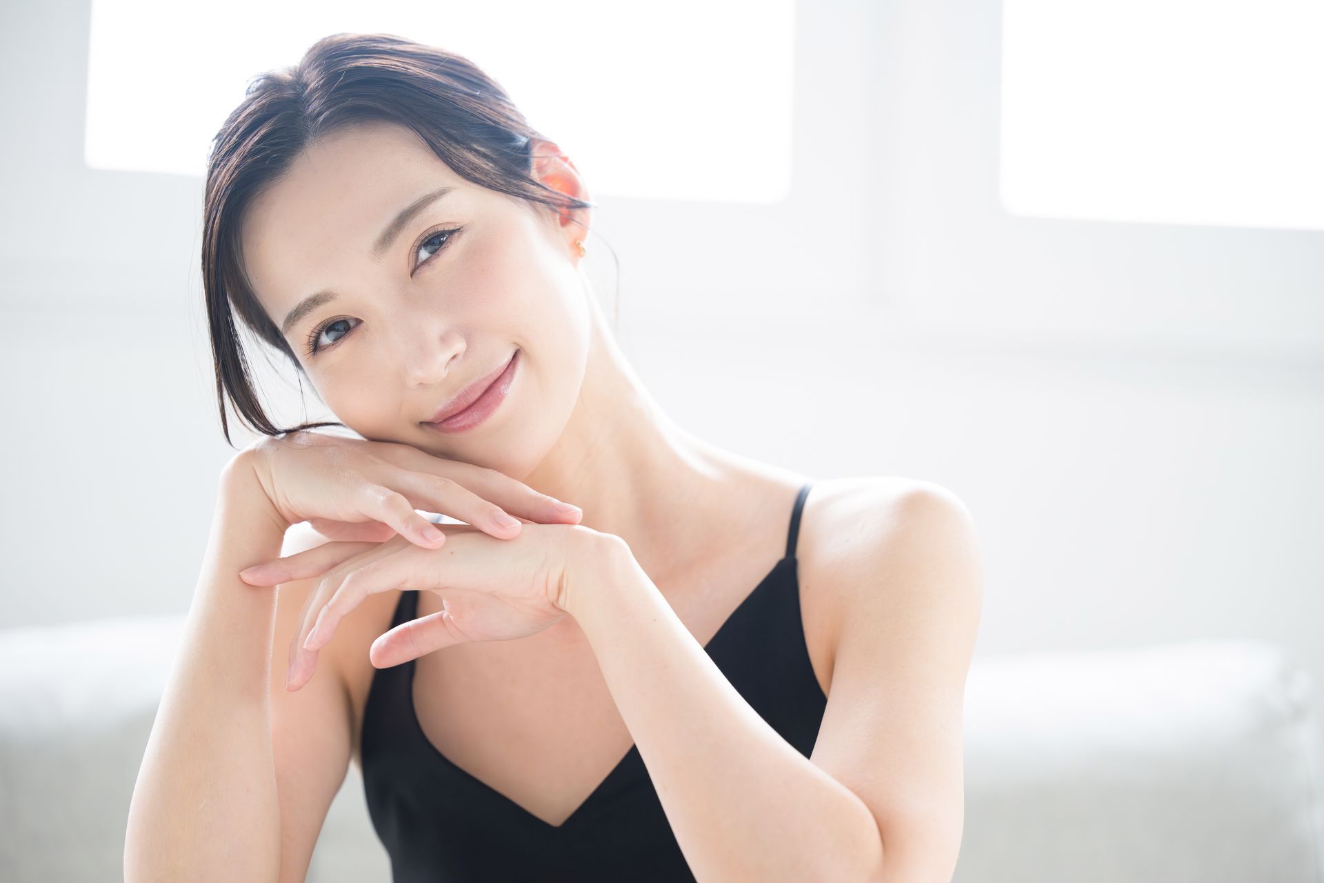 Woman in black top resting chin on hands, smiling, in bright room.