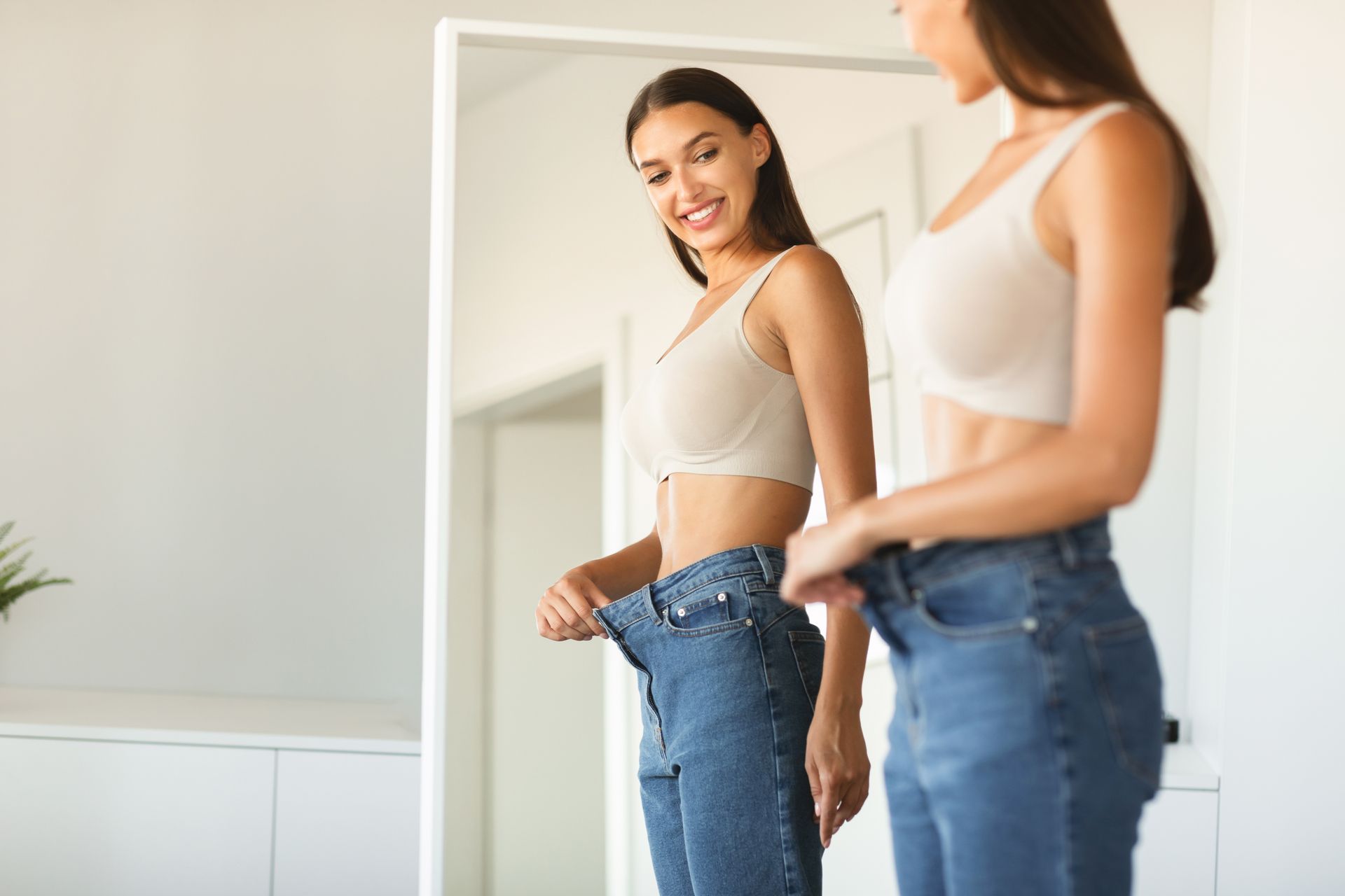Woman smiling, wearing loose jeans, looking at herself in a mirror, implying weight loss.