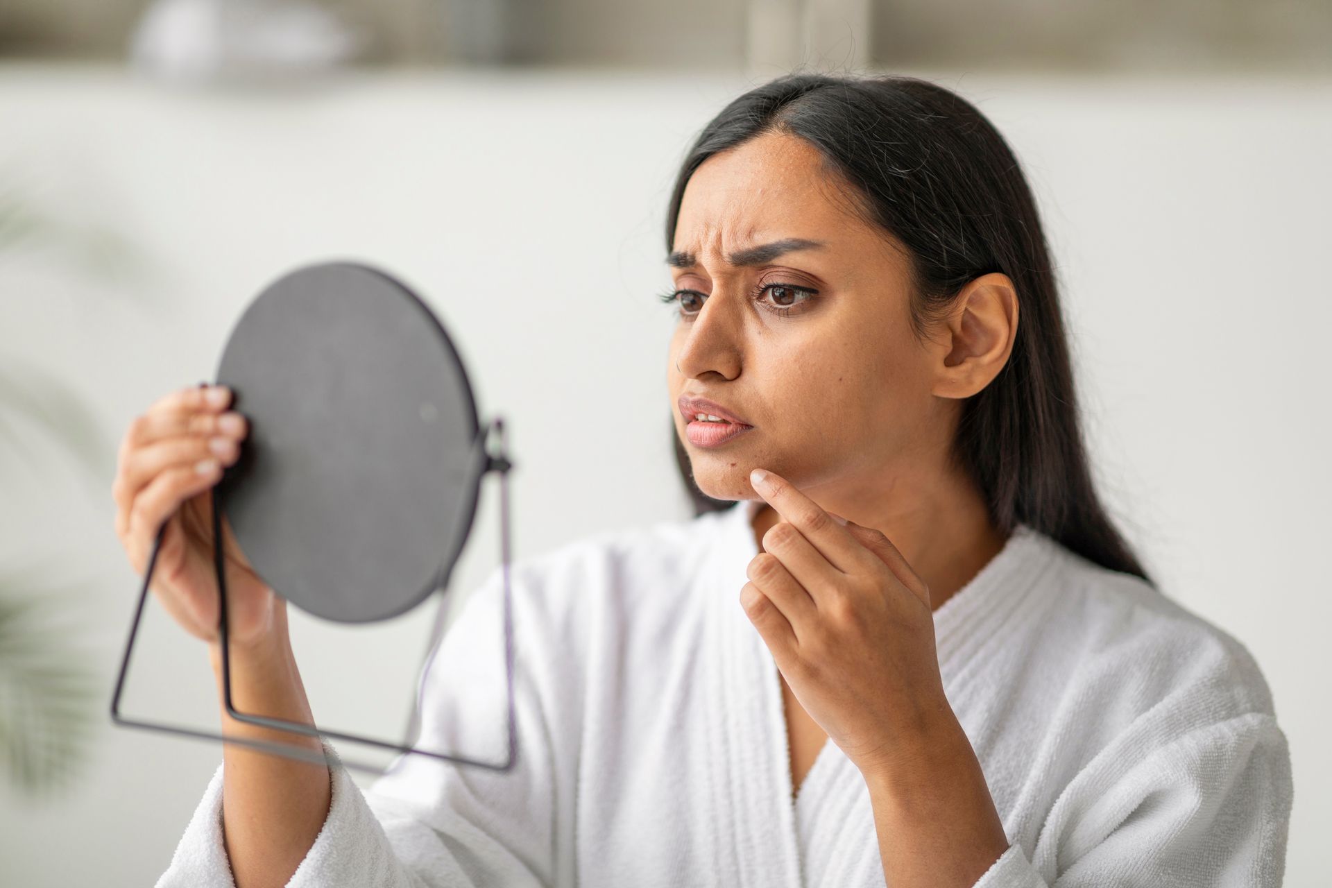 Woman examining her face in a handheld mirror, looking concerned in a bright room