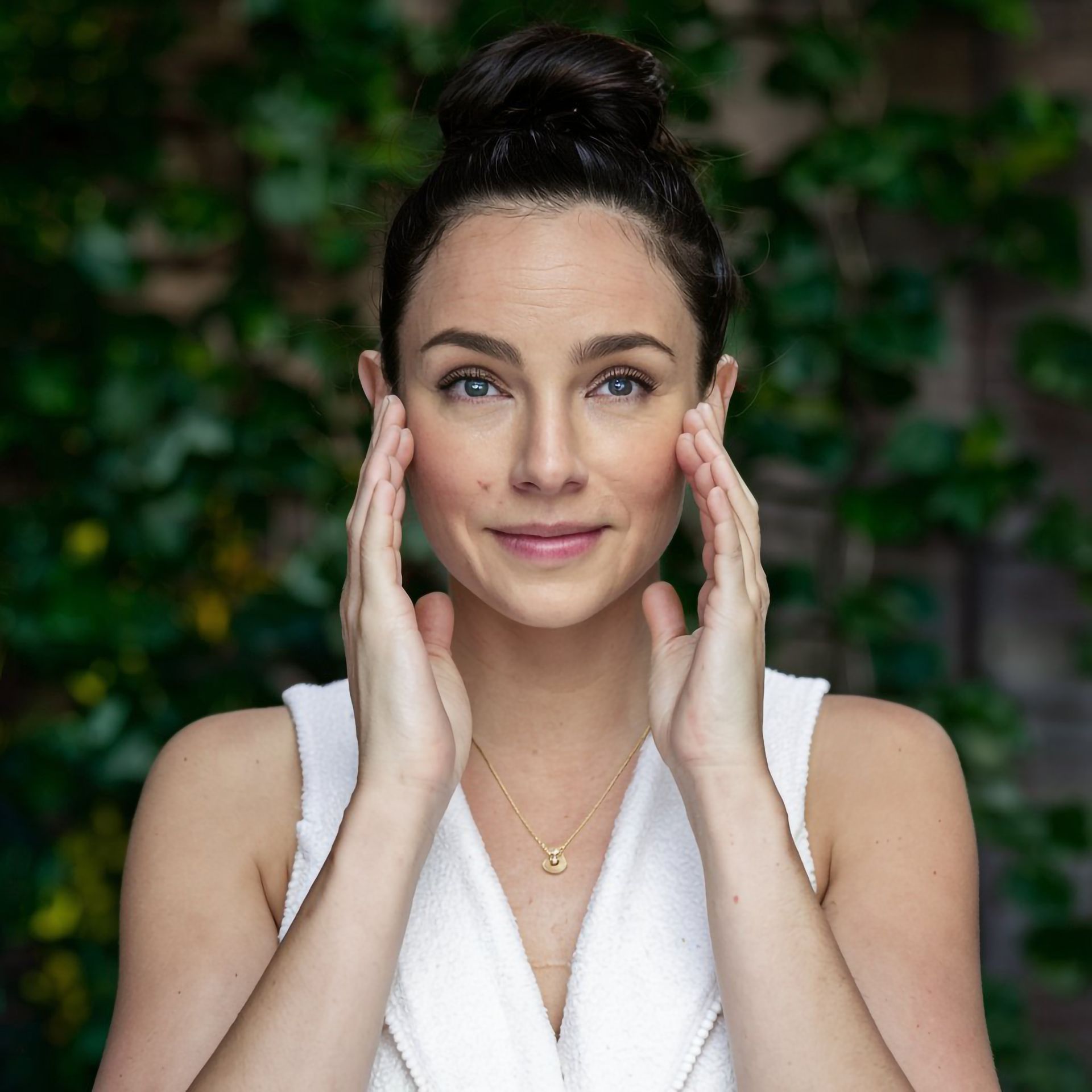 Woman in white robe, hands on face, looking at the camera, with a bun, in front of greenery.