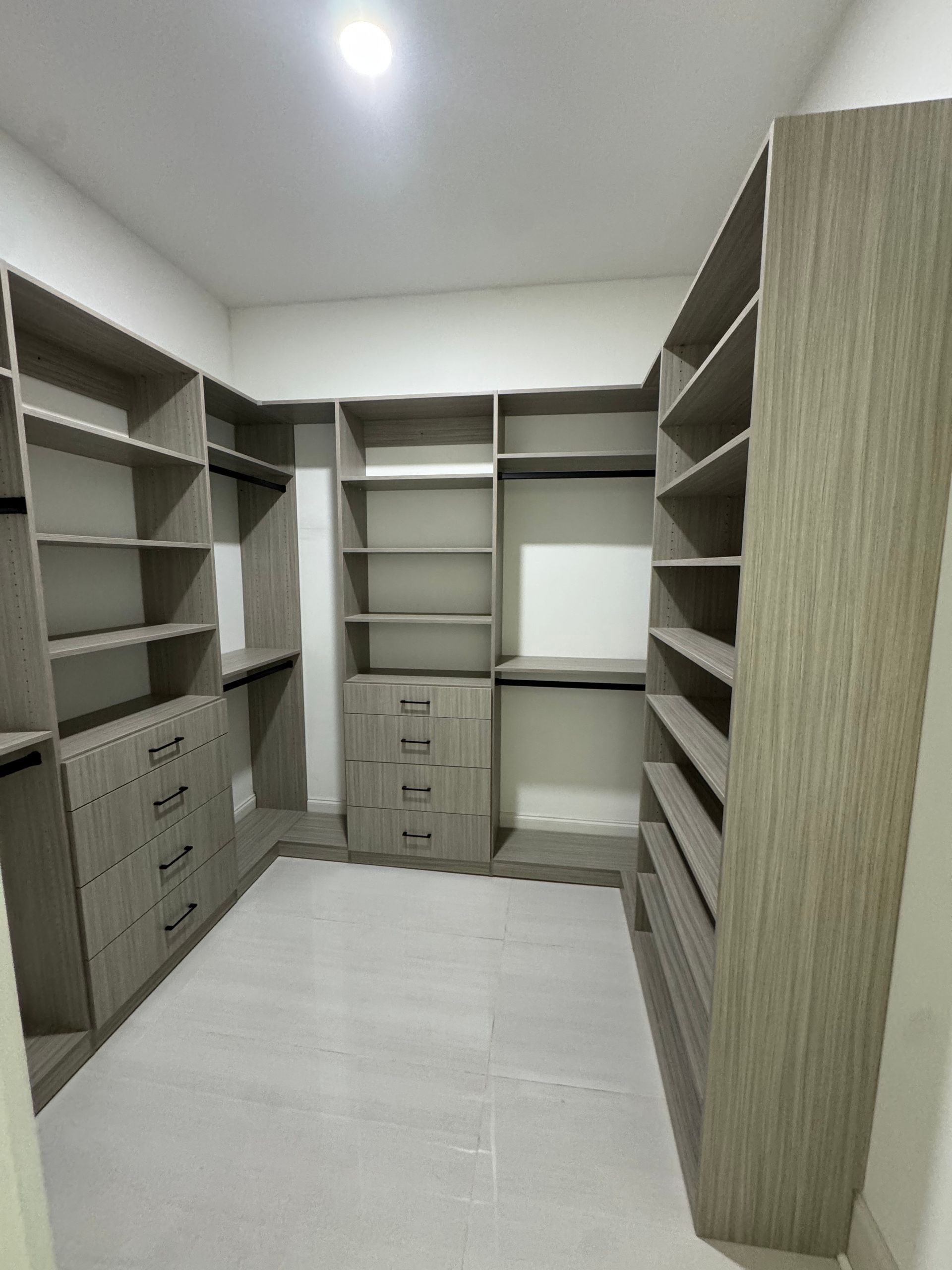 Empty walk-in closet with built-in shelving and drawers, light wood-grain finish. Grey tile floor and white walls.