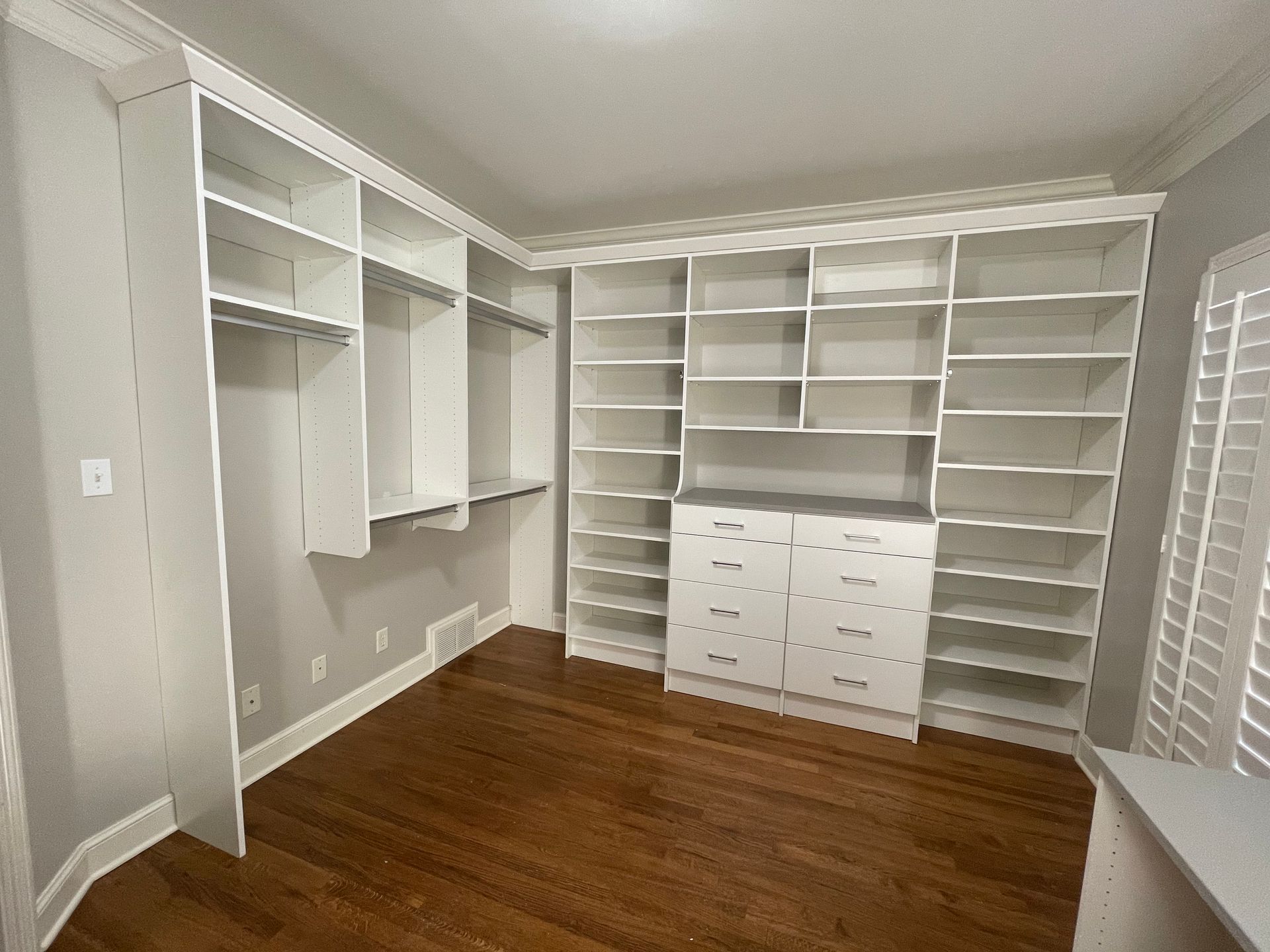 Empty white built-in closet with shelves, drawers, and hanging rods against gray walls and hardwood floor.