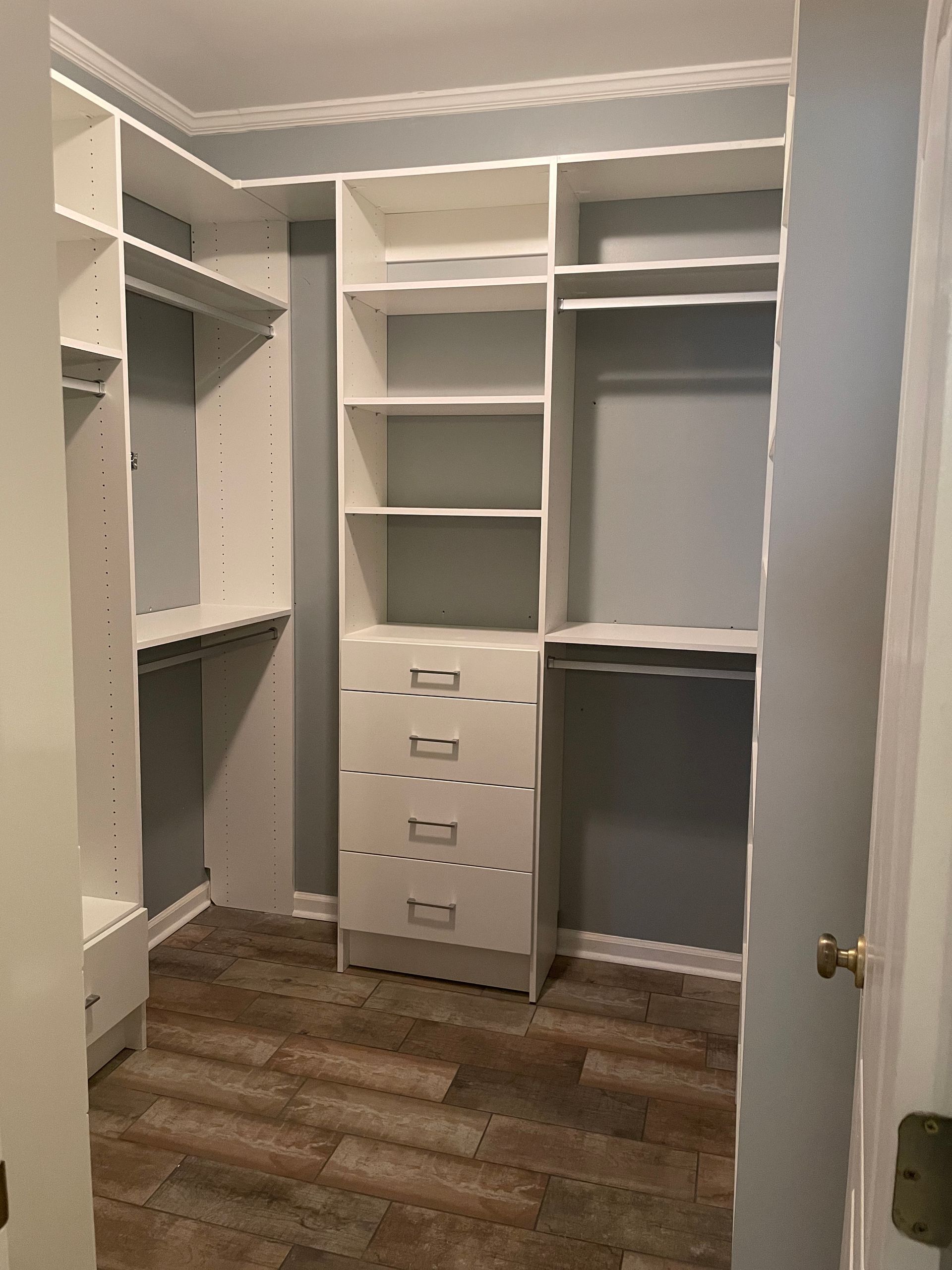 White closet organizers in a walk-in closet with shelves, drawers, and hanging rods. Gray walls, wood-look floor.