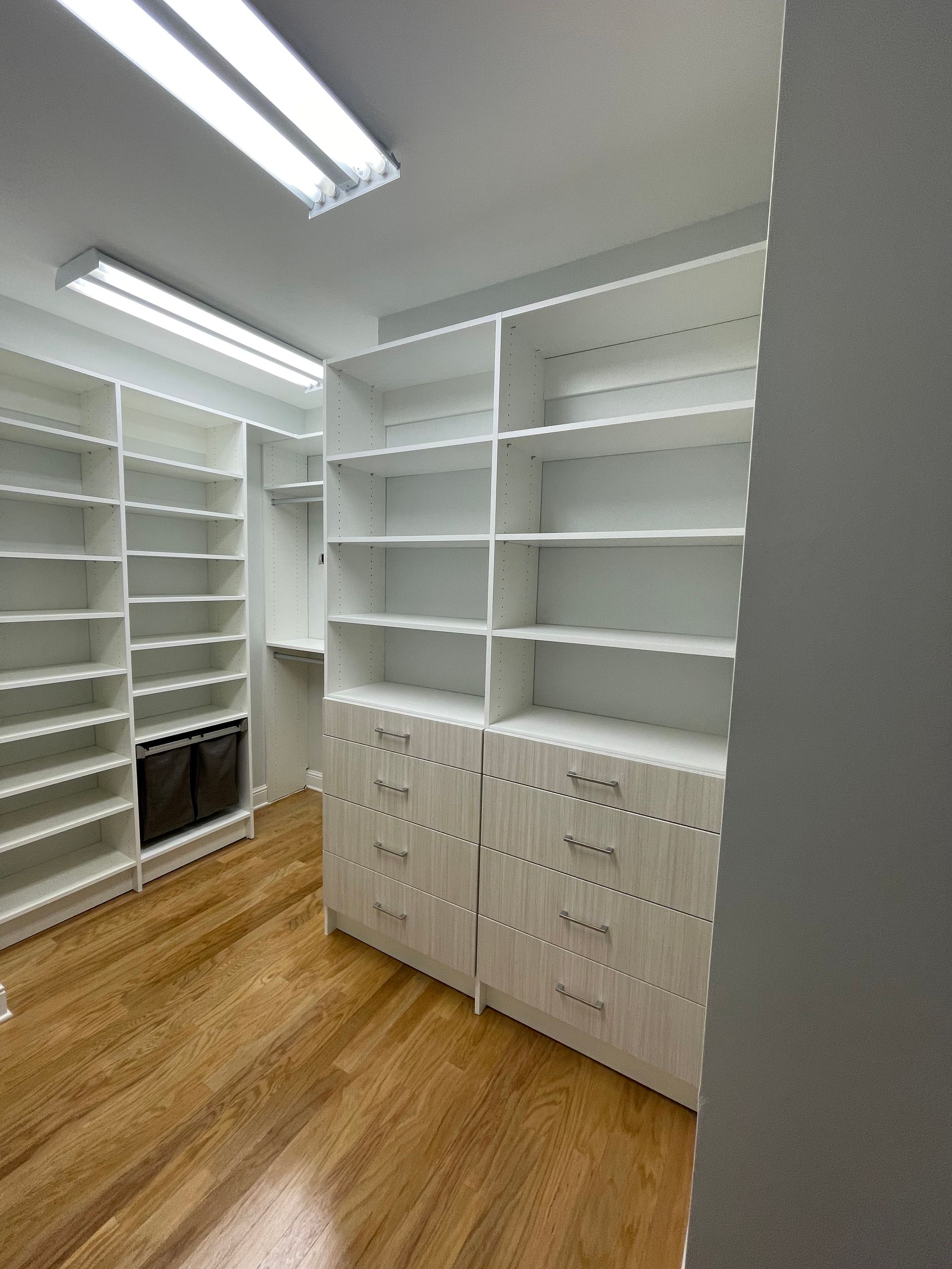 Walk-in closet with white shelving and drawers.  Wooden floors, overhead lighting.