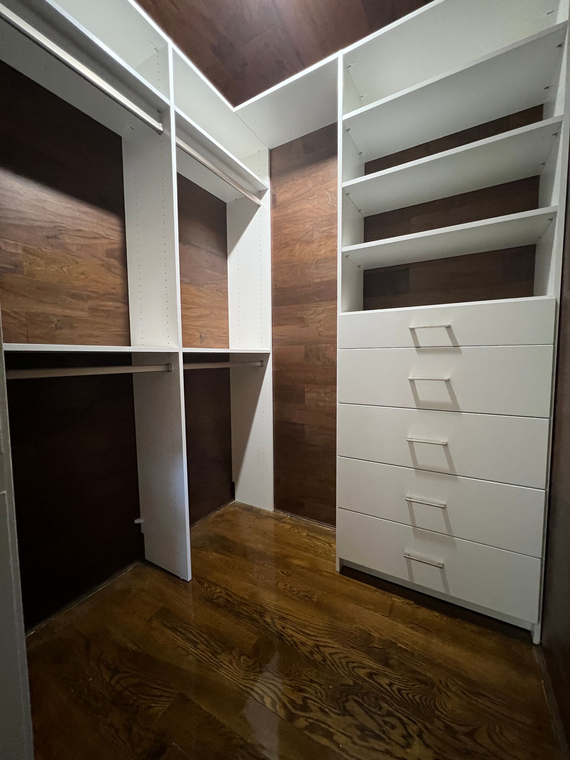 Walk-in closet with white shelves and drawers against wood-paneled walls and floor.