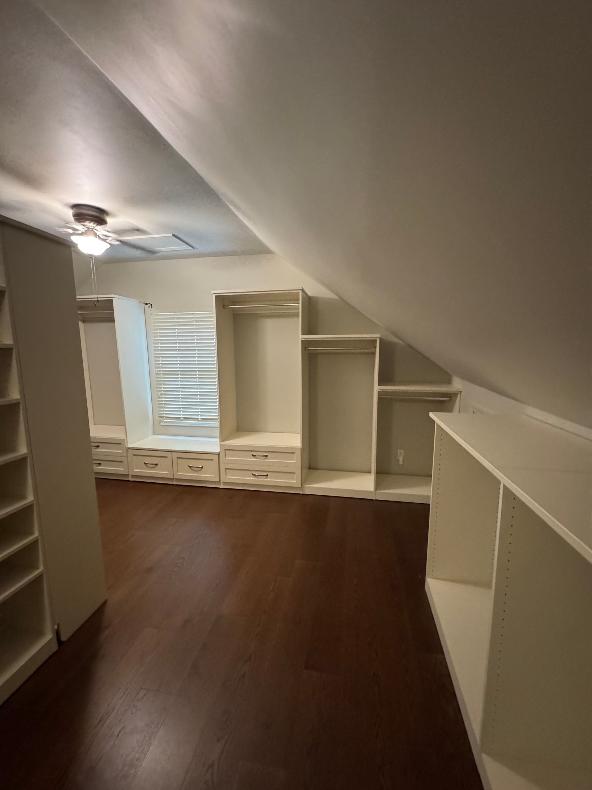 Walk-in closet with white built-in storage on dark wood floor, under angled ceiling with a window.