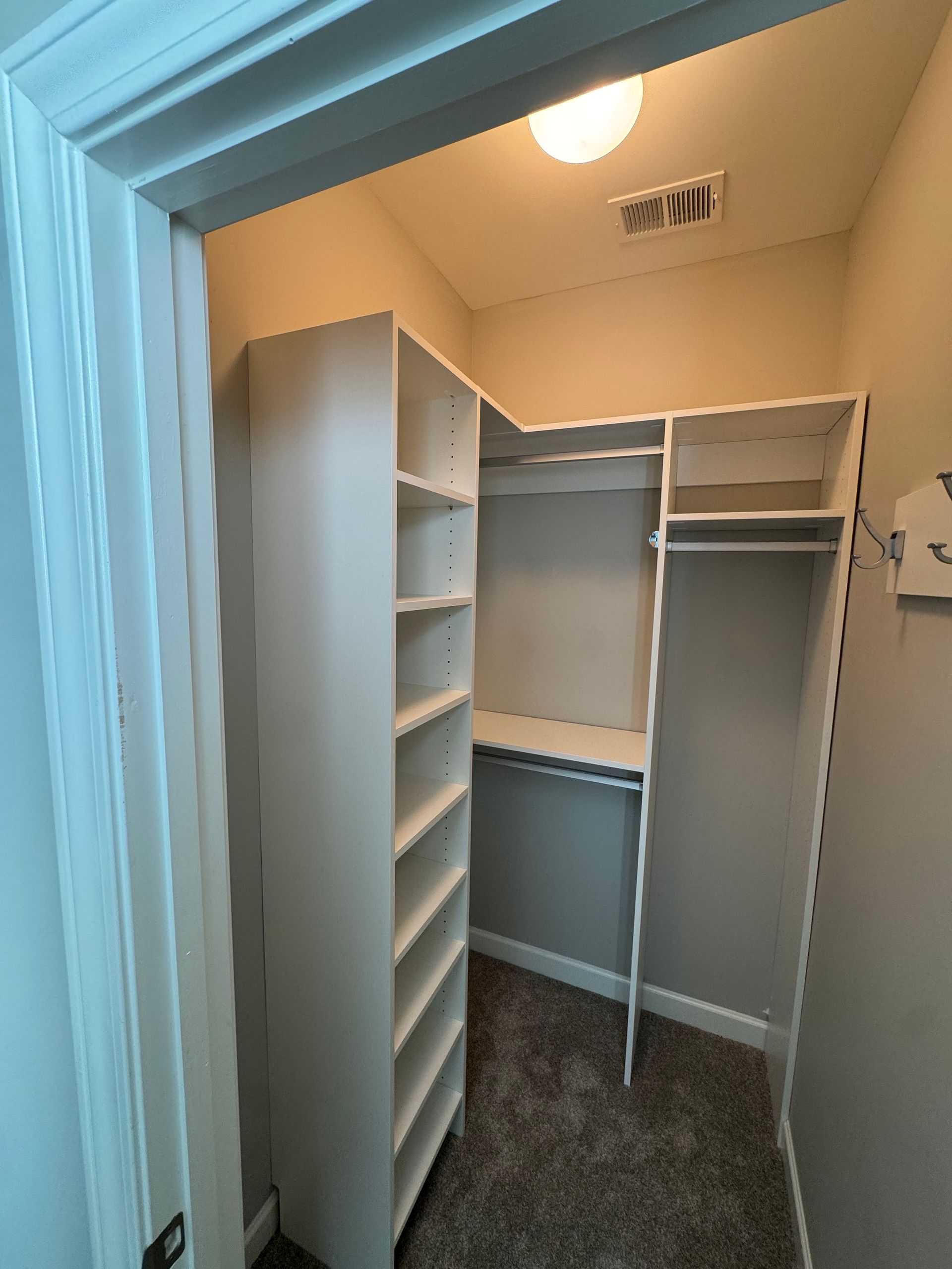 Small walk-in closet with built-in shelves and hanging rods; neutral colors, carpet flooring.