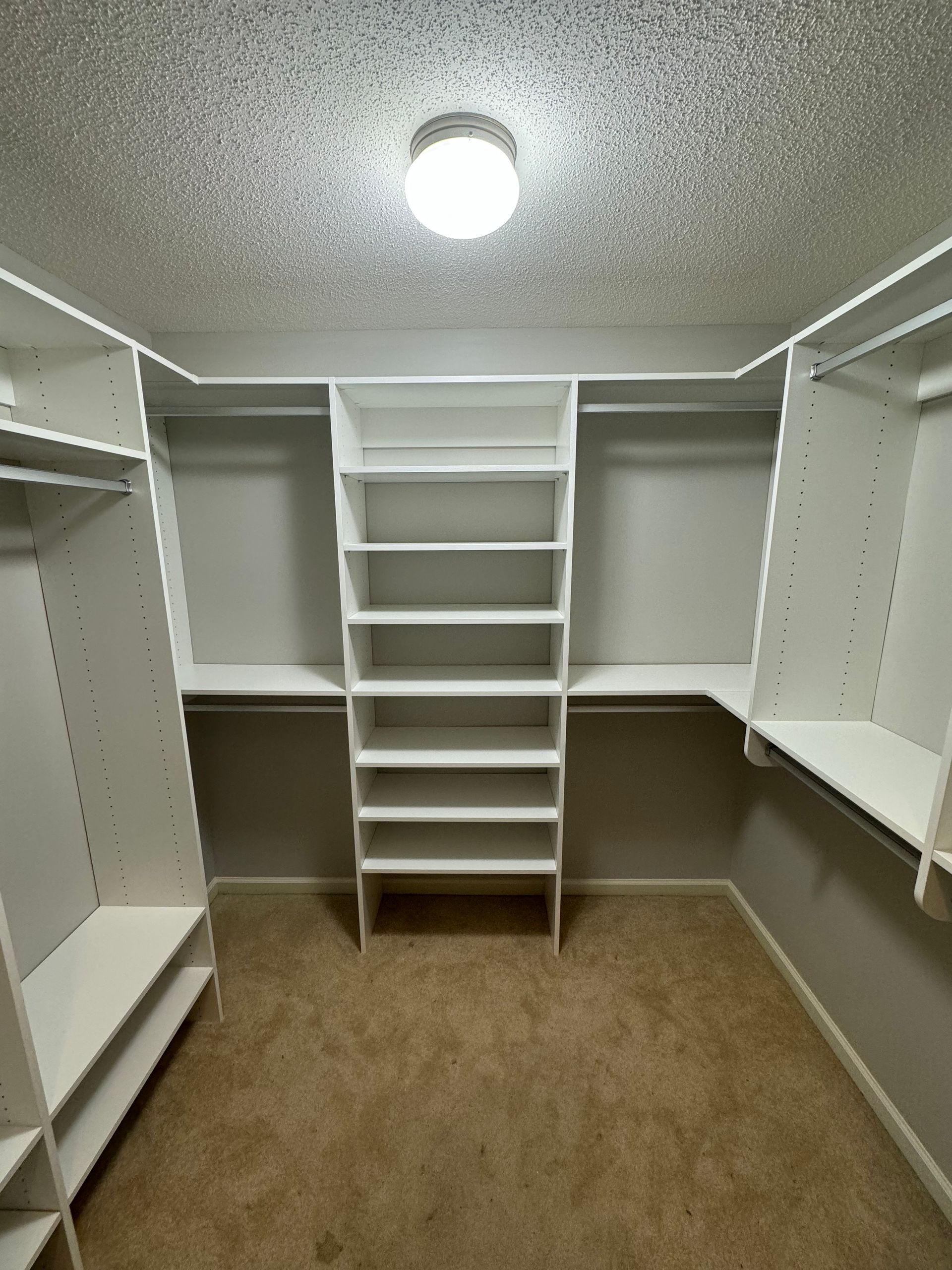 Empty walk-in closet with white shelving and hanging rods. Tan carpet, recessed lighting, and a neutral color on the back.