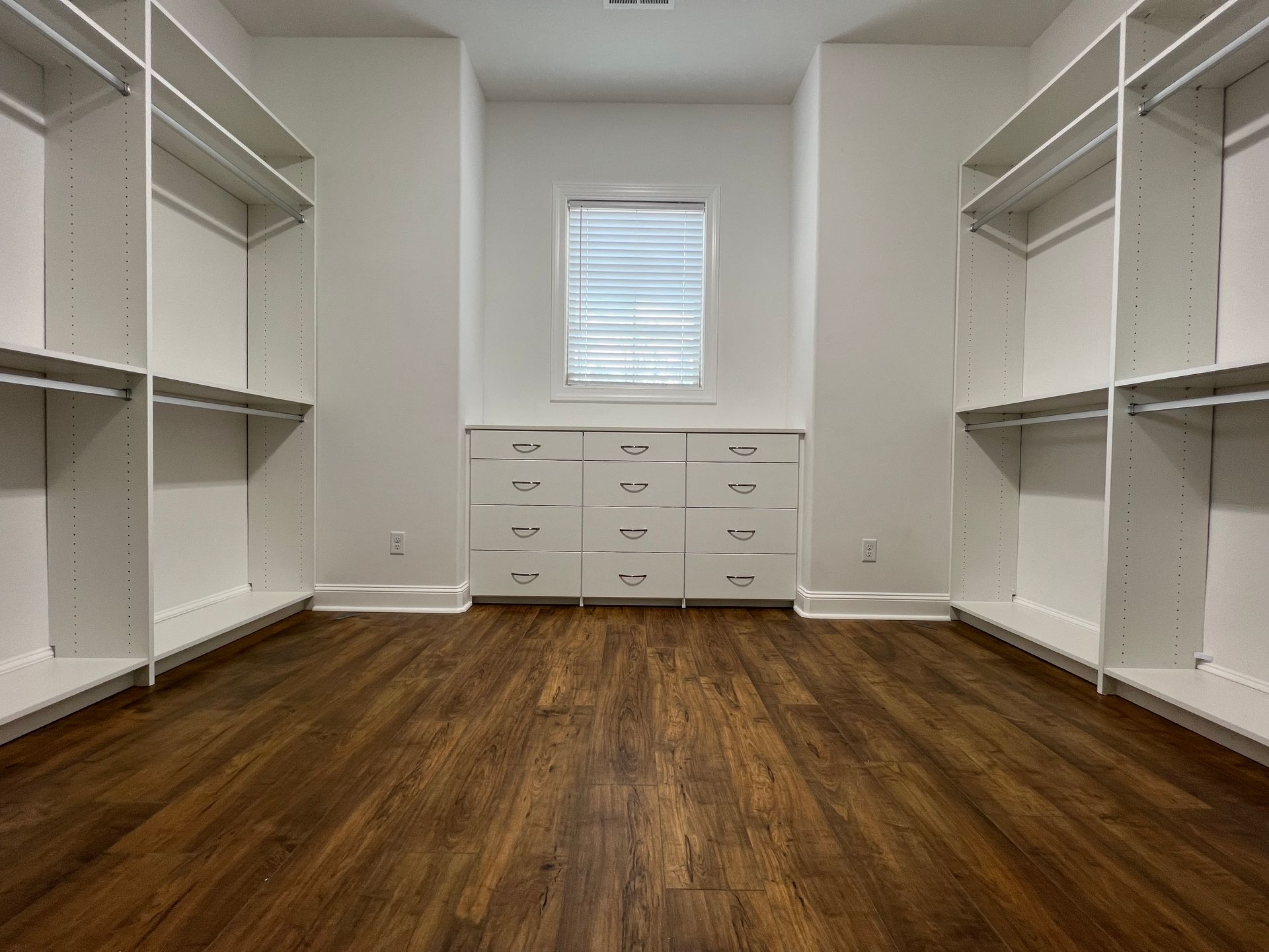 Empty, white-walled walk-in closet with shelves and drawers. Dark wood floor, window with blinds.
