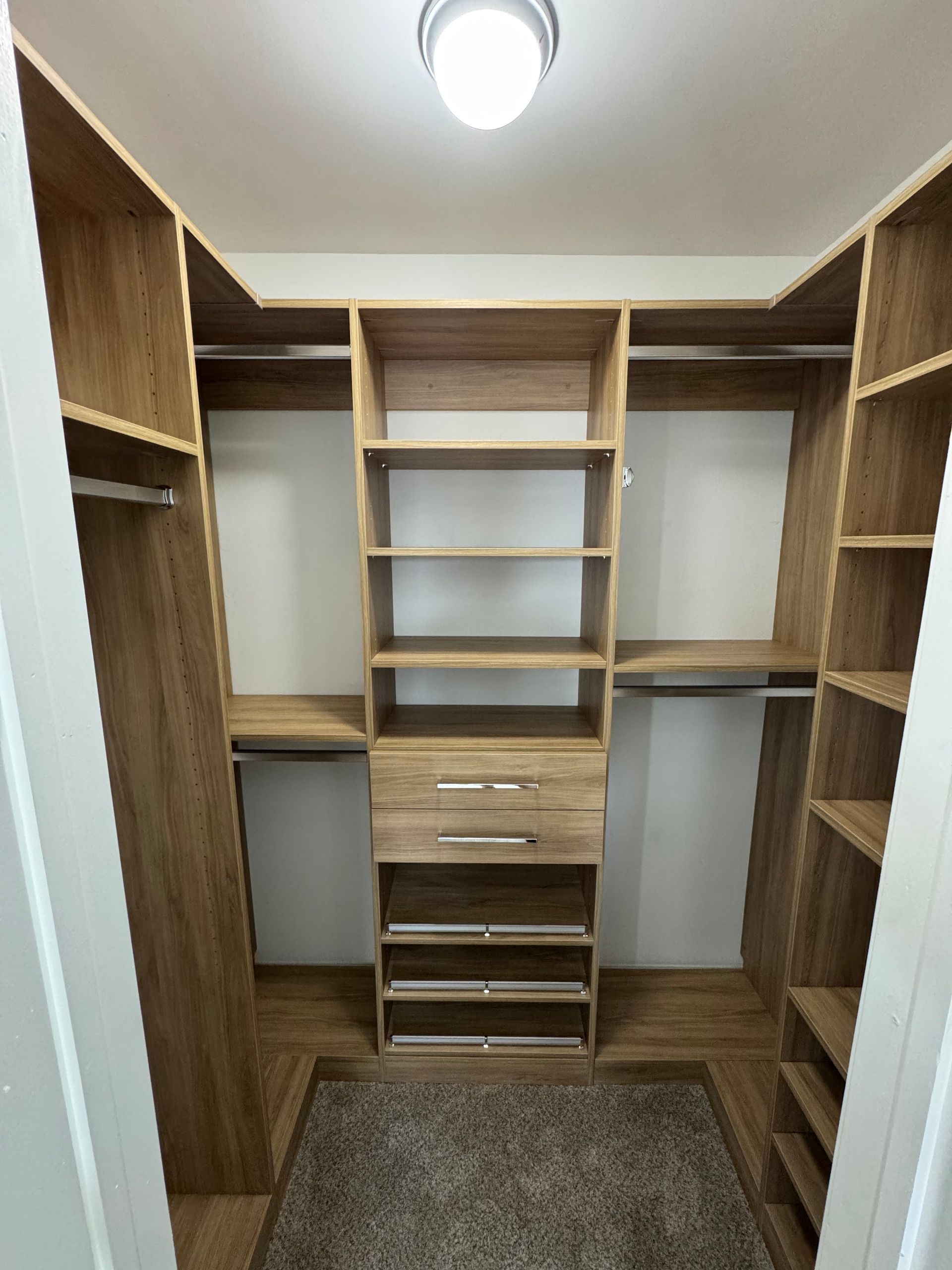 U-shaped wooden closet interior with shelves, drawers, and hanging rods. Light-colored wood and neutral walls.