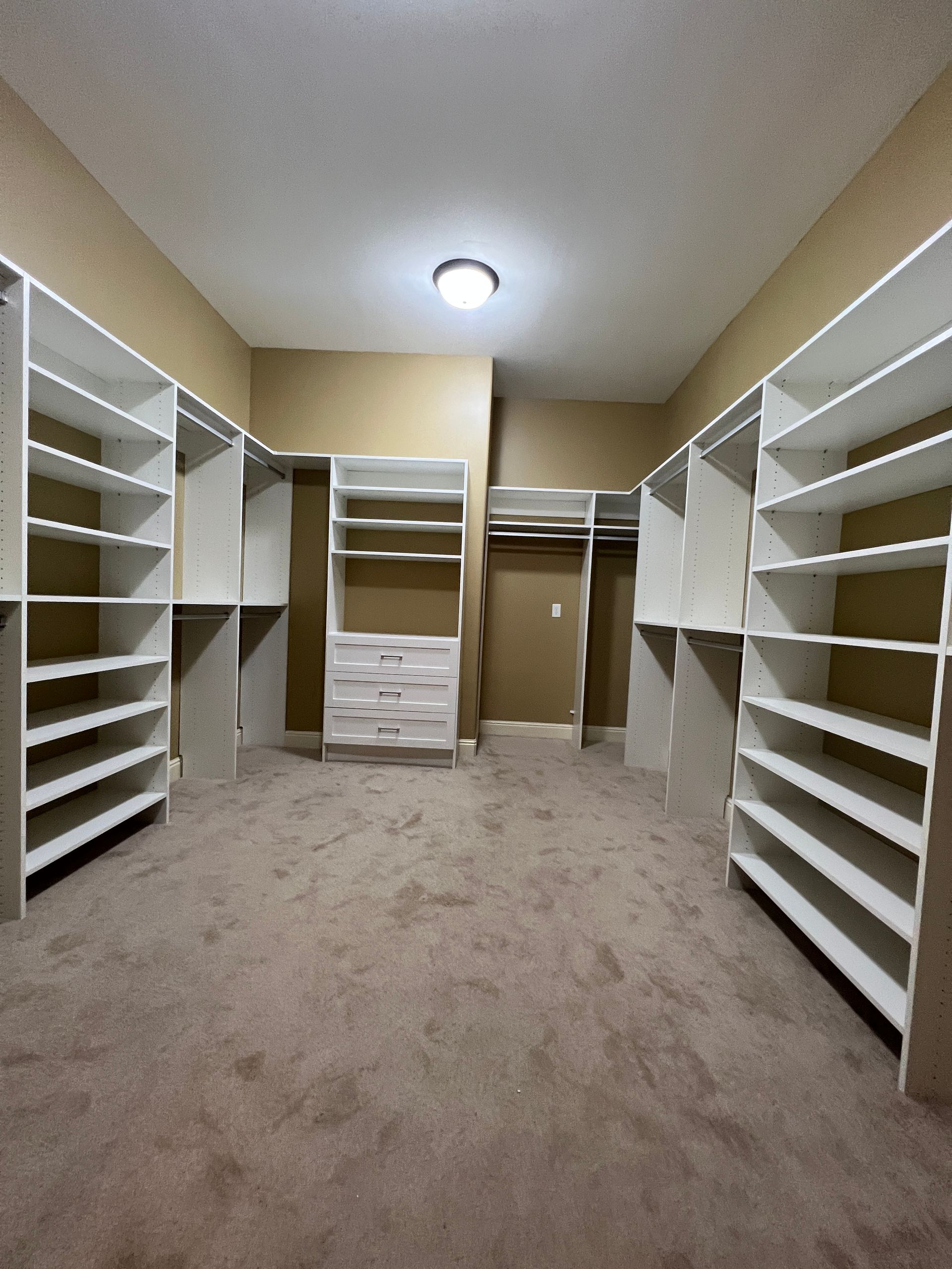 Empty walk-in closet with white shelving on either side of a tan carpeted floor. The walls are a beige color.