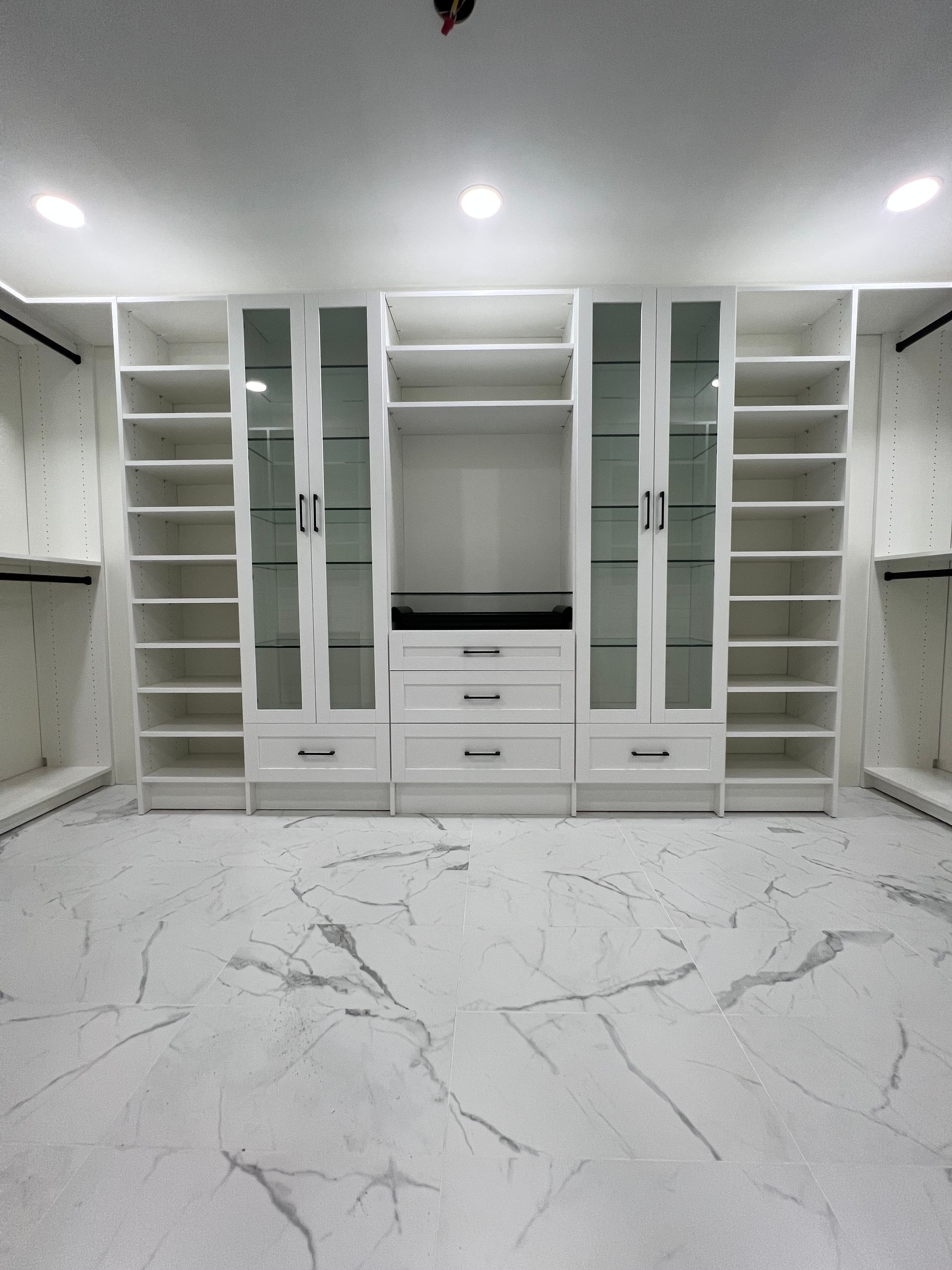 White custom walk-in closet with shelves, glass doors, and drawers, set against a marble floor.