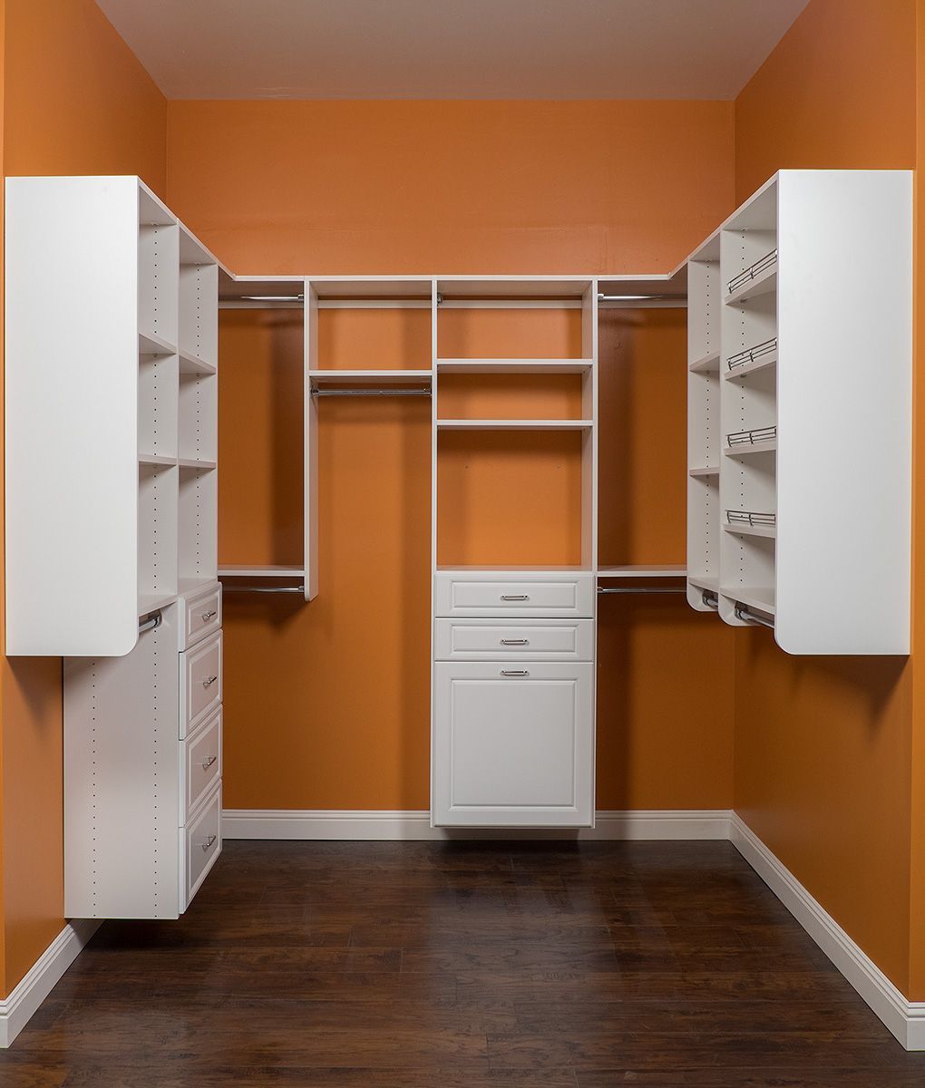 Empty walk-in closet with white shelves, drawers, and rods against orange walls. Dark wood floor.