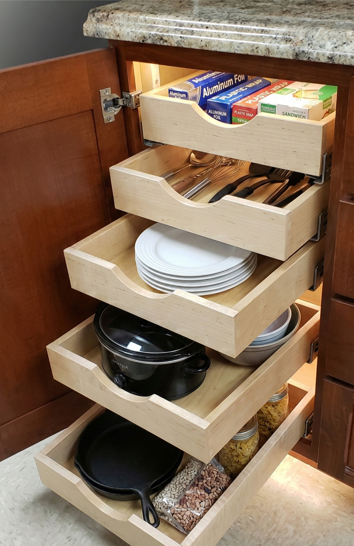 Pull-out kitchen cabinet with various contents: foil boxes, silverware, plates, pots, and jars.