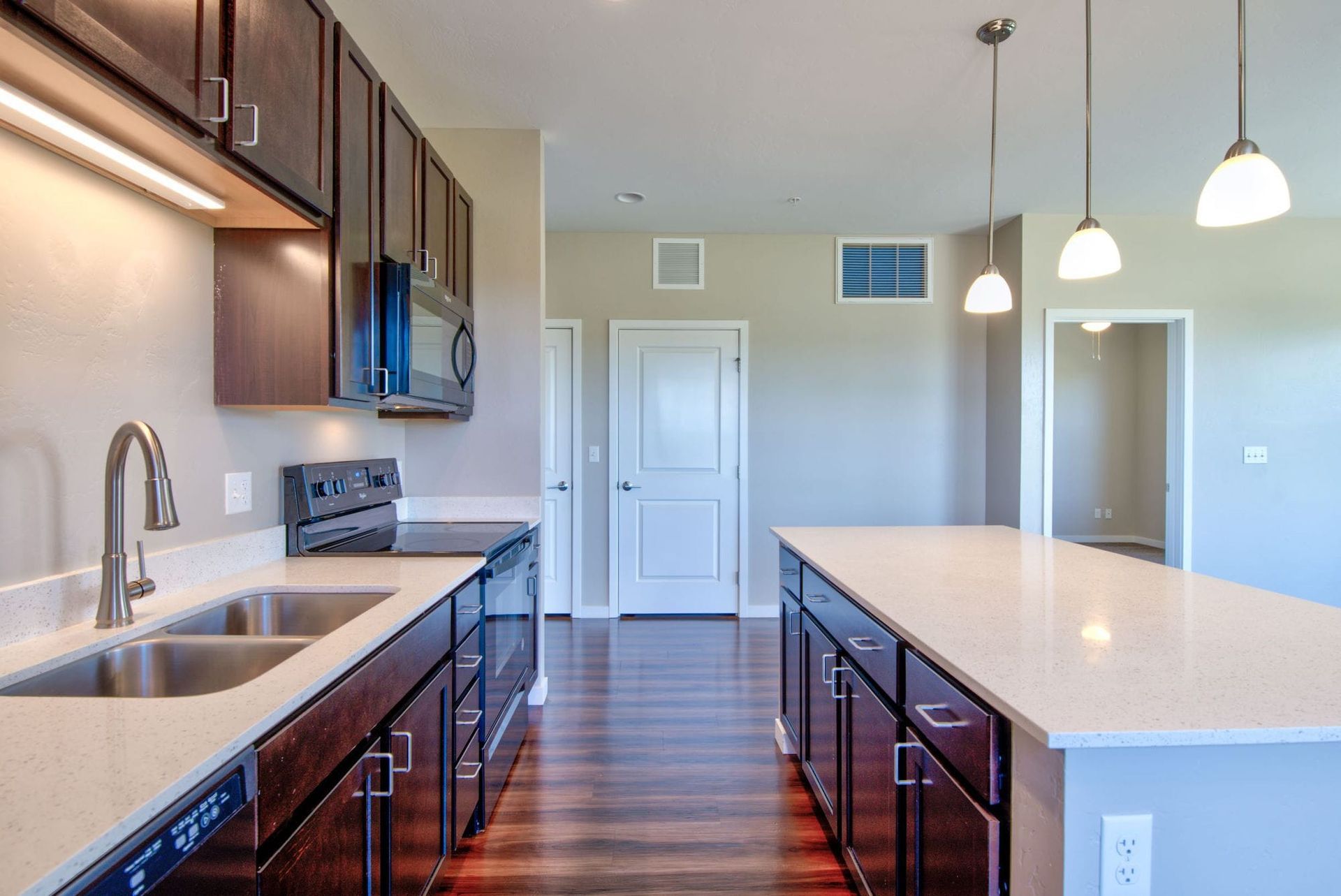 People working at a large wooden table in a bright office; two using laptops, one writing, plants and supplies on the table.