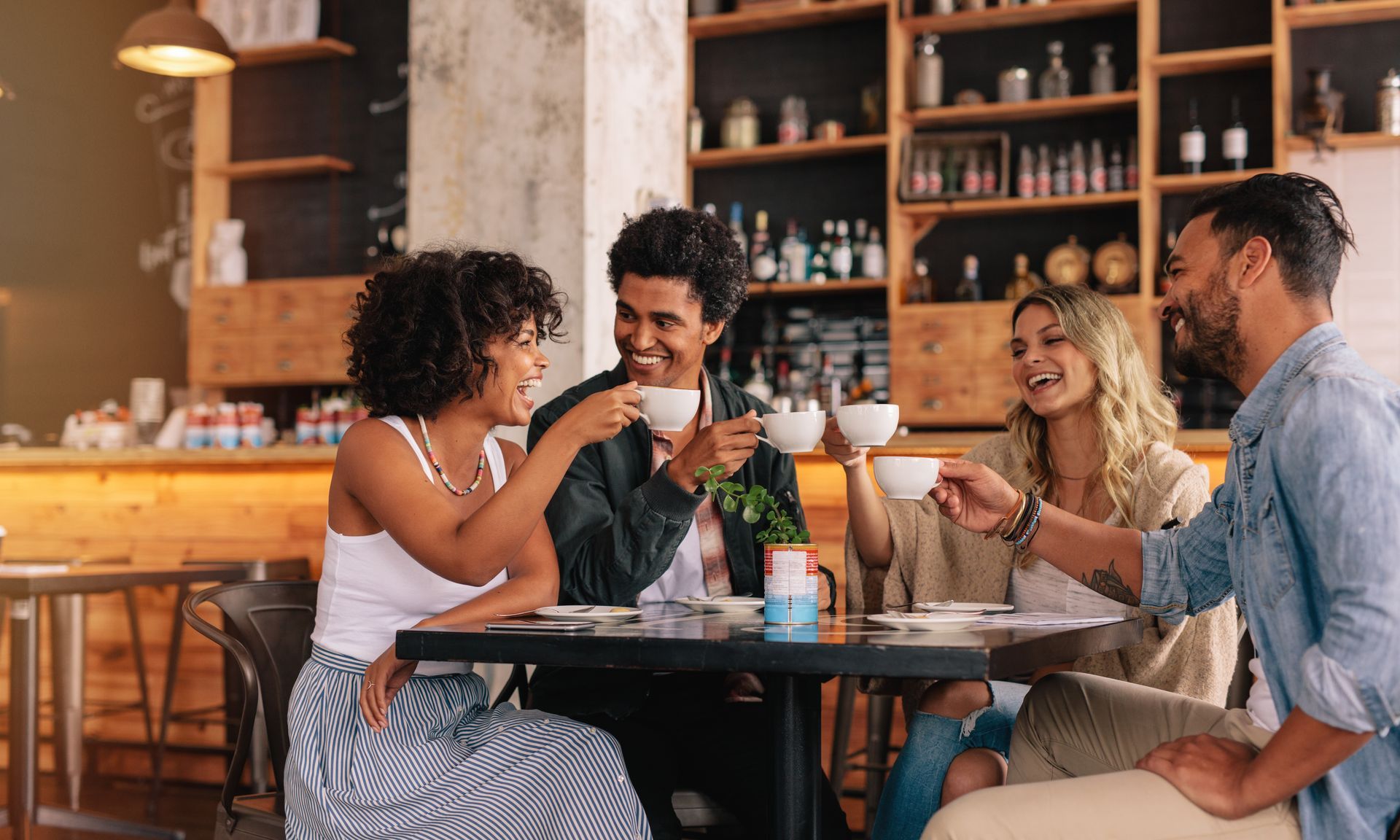 Four friends at a cafe, raising cups in a toast. Smiling, light-filled setting with wood accents near Tradewinds Apartments