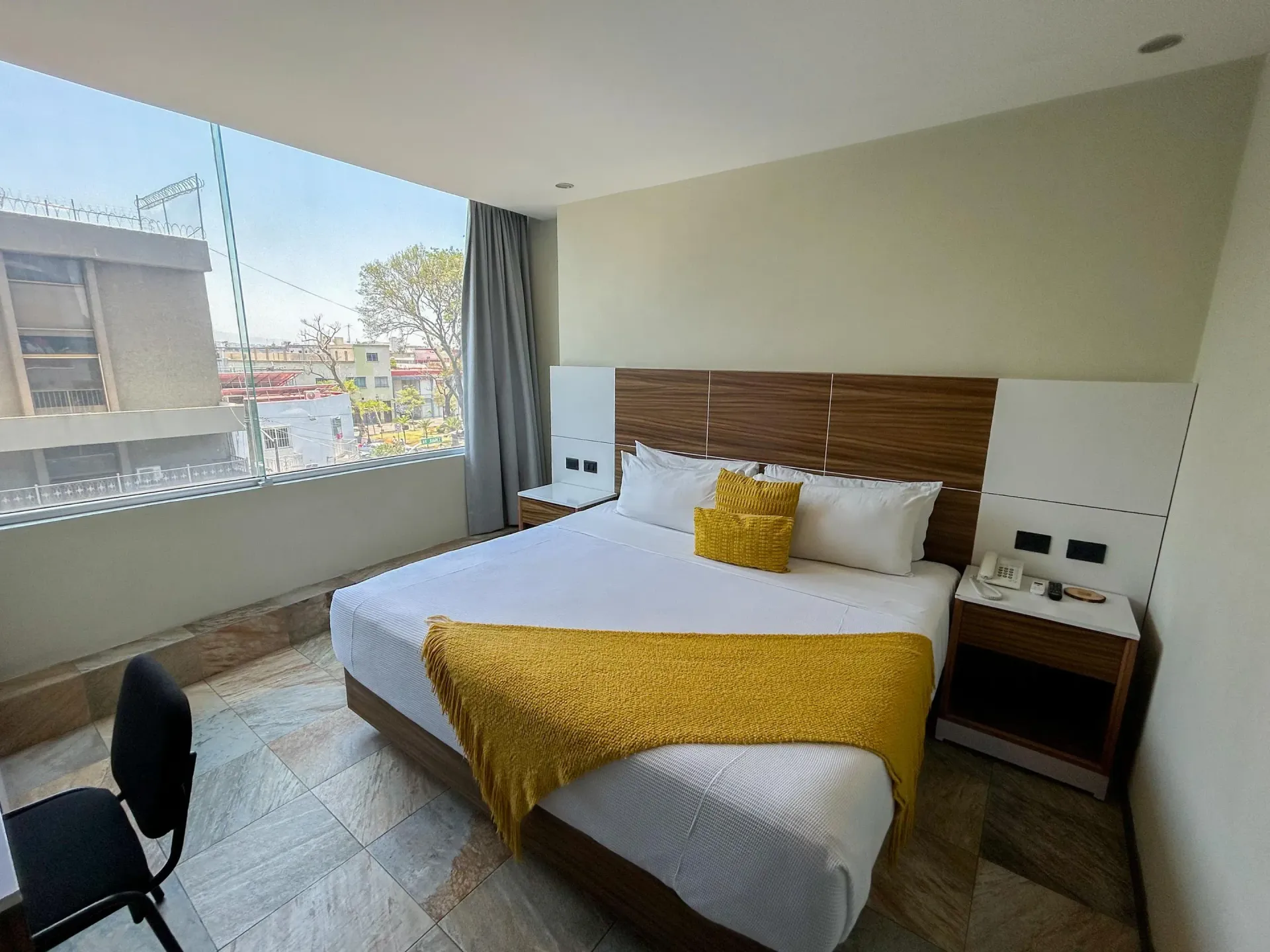 Habitación de hotel luminosa con cama, manta amarilla, silla de escritorio y gran ventana con vistas.