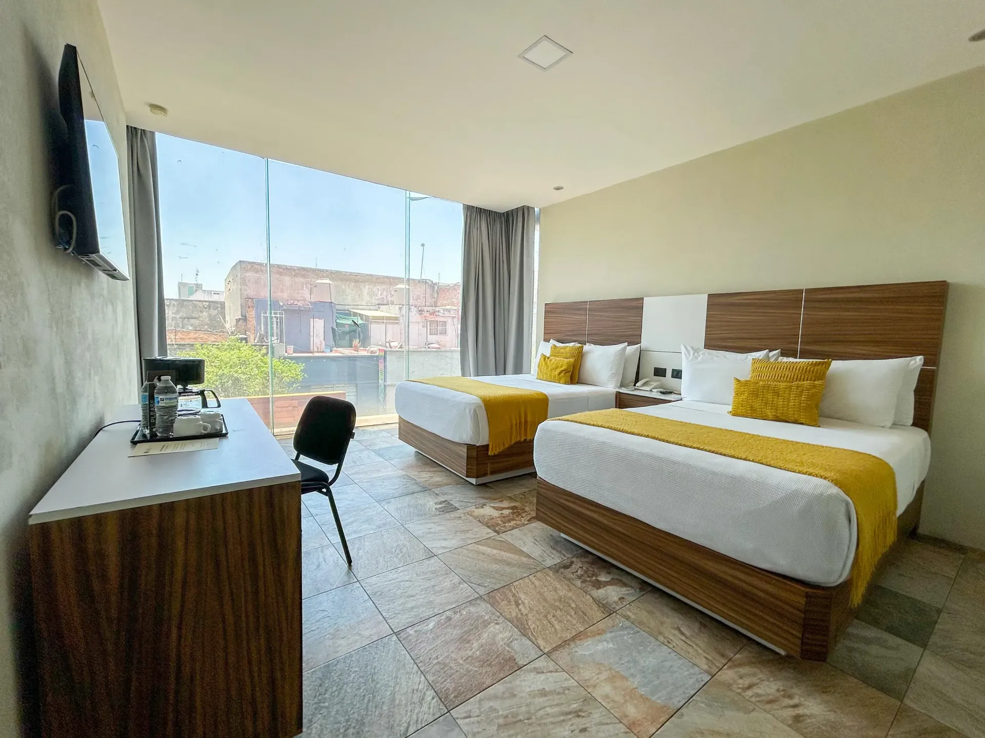 Habitación de hotel con dos camas, escritorio, silla, ventana grande y vista a la ciudad.