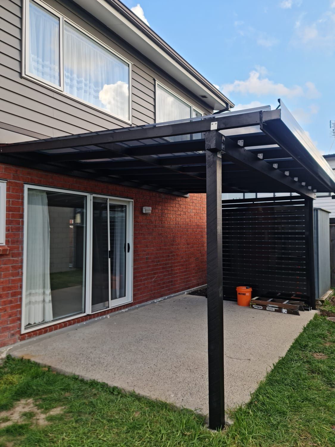 Black pergola over a concrete patio next to a brick house and sliding glass door.