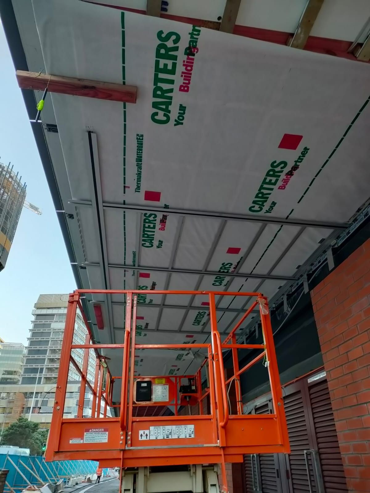 Orange lift platform supporting workers installing building wrap labeled 