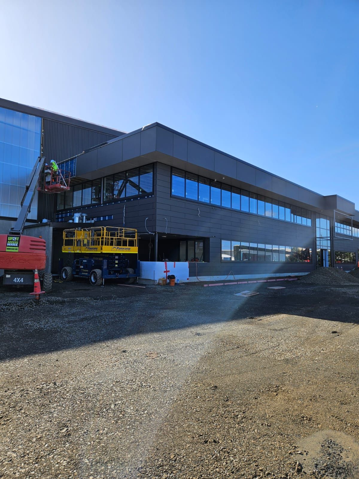 Construction of a two-story building on a gravel lot; workers in lift completing exterior work on a sunny day.