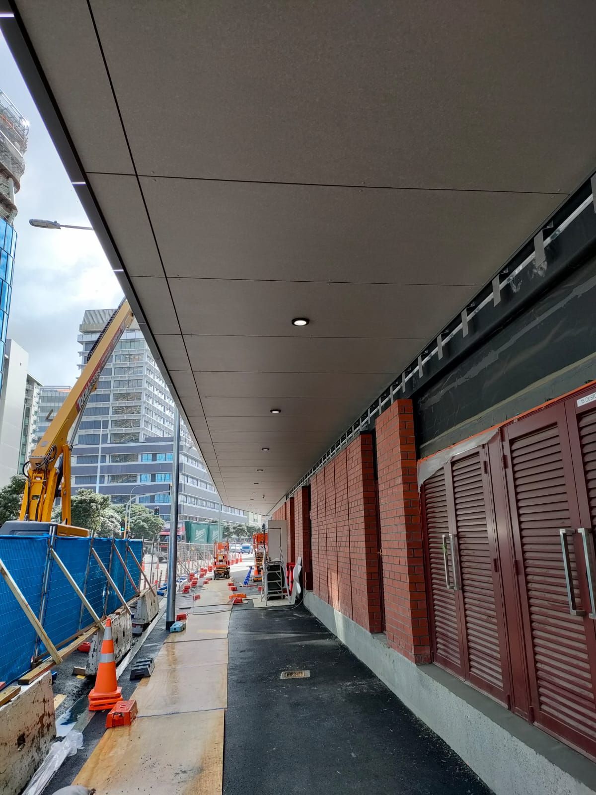 Sidewalk under a covered walkway. Brick wall with closed shutters on right, construction equipment in background.