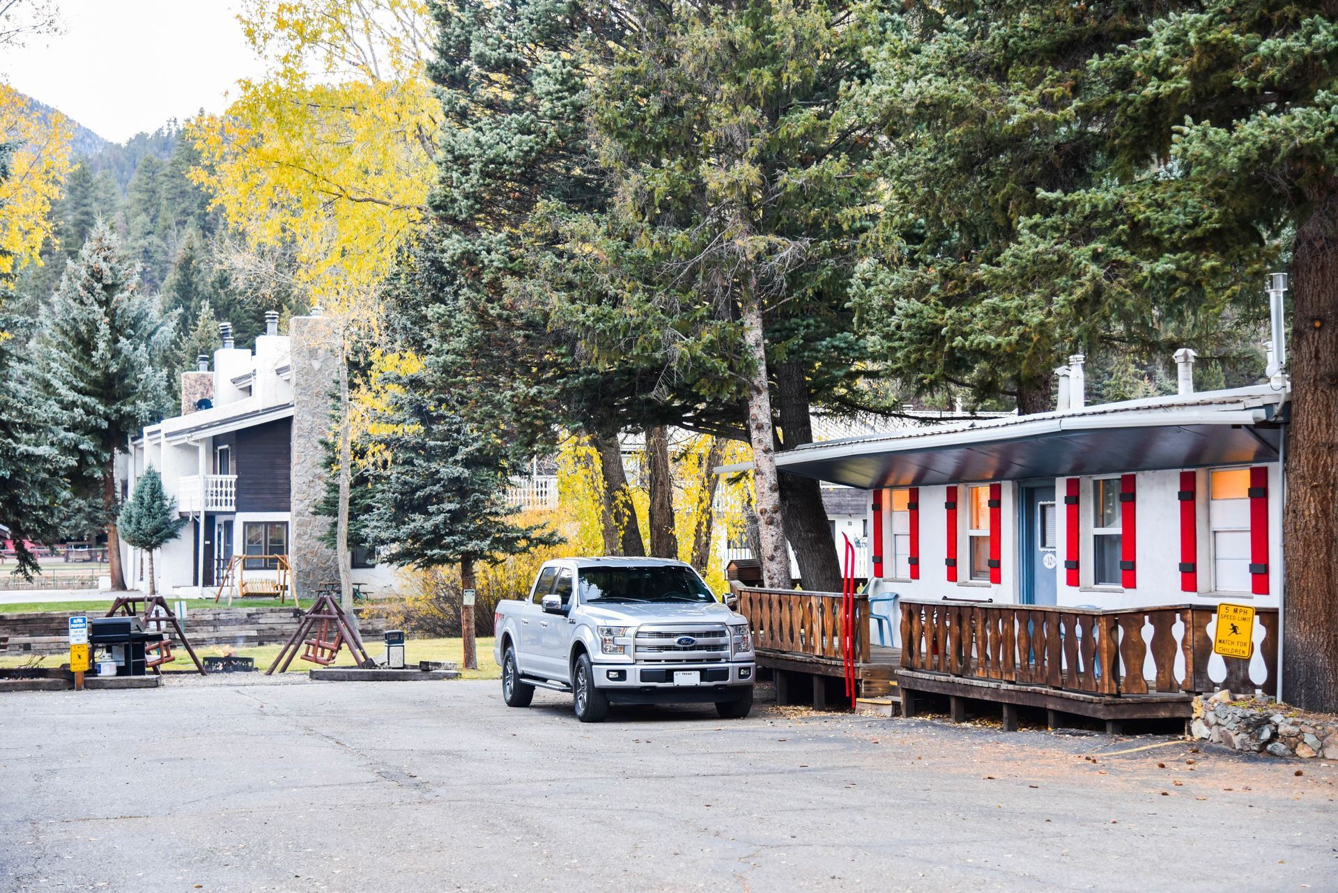 A silver pickup truck parked in front of a white cabin with red shutters, trees, and a mountain backdrop.