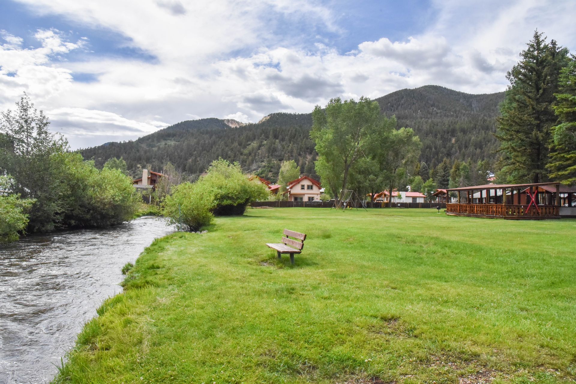 A grassy park by a river, with a bench, trees, and mountains in the background under a cloudy sky.