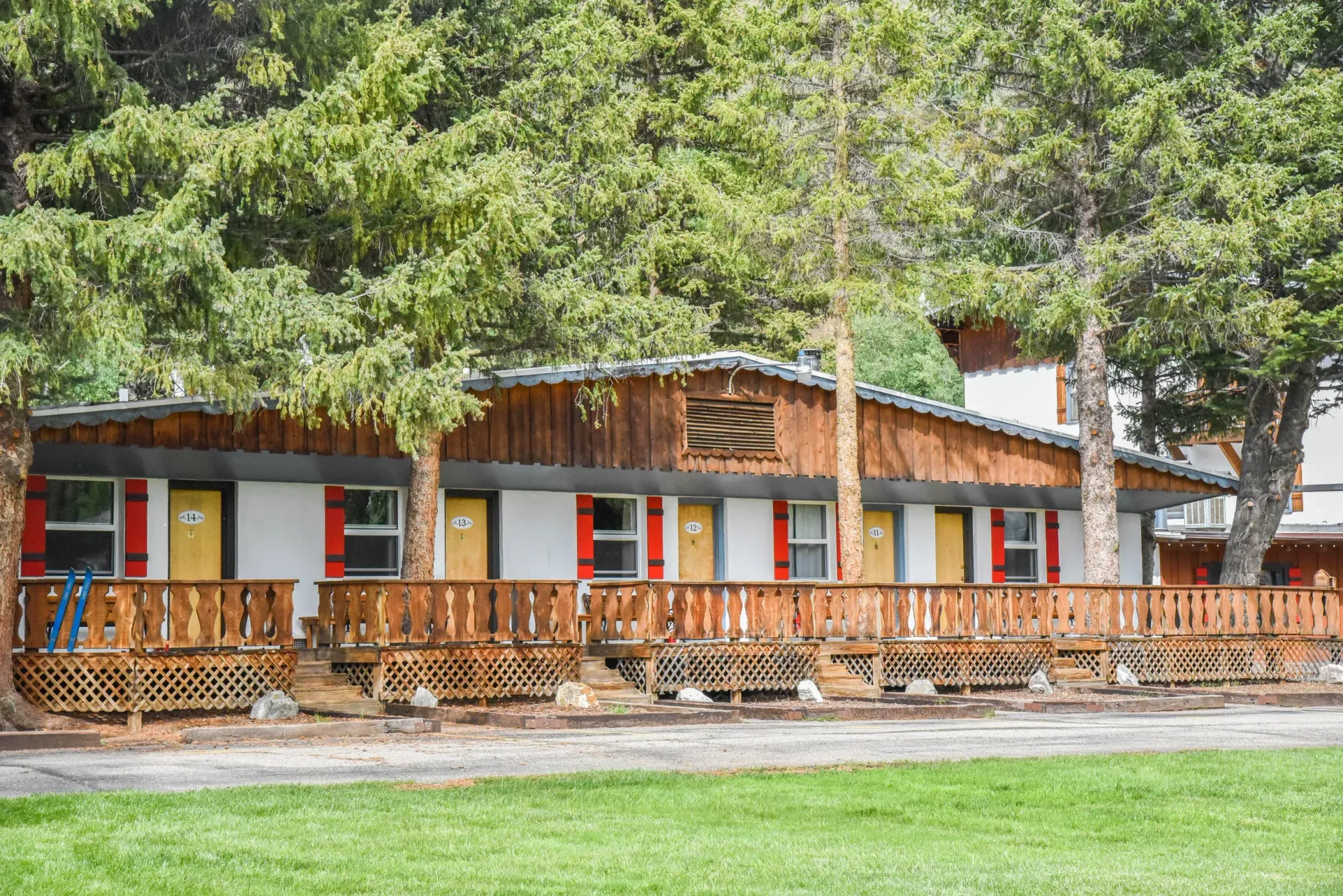 A motel with brown wood trim, red shutters, and yellow doors sits behind a wooden deck, with trees in the background.