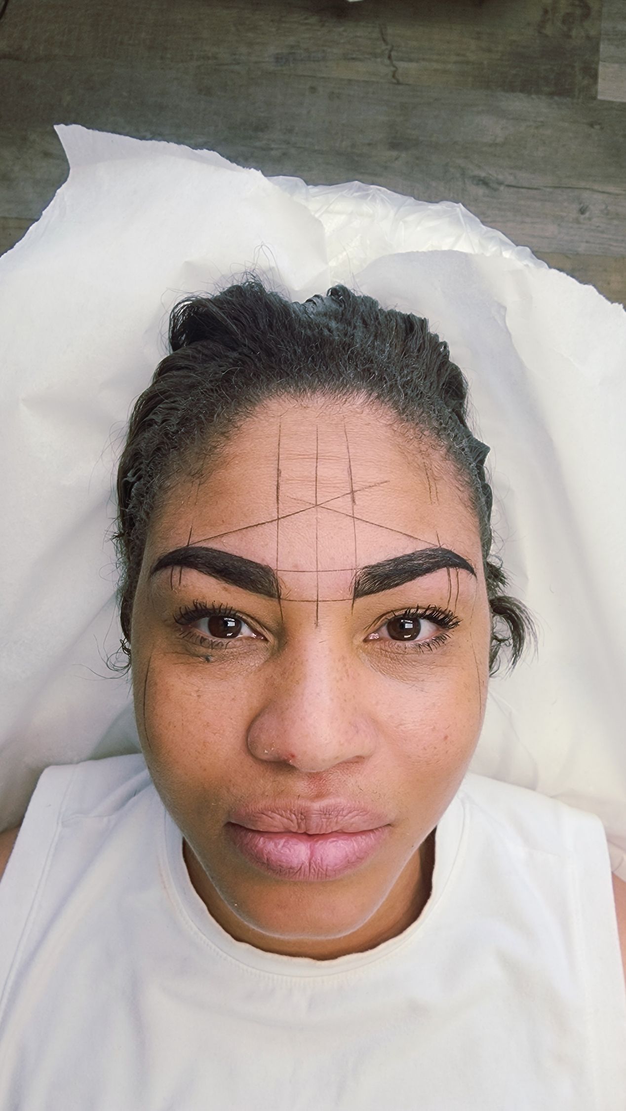 Woman with marked eyebrows, prepared for a cosmetic procedure. Fine lines outline eyebrow shape, on a white surface.