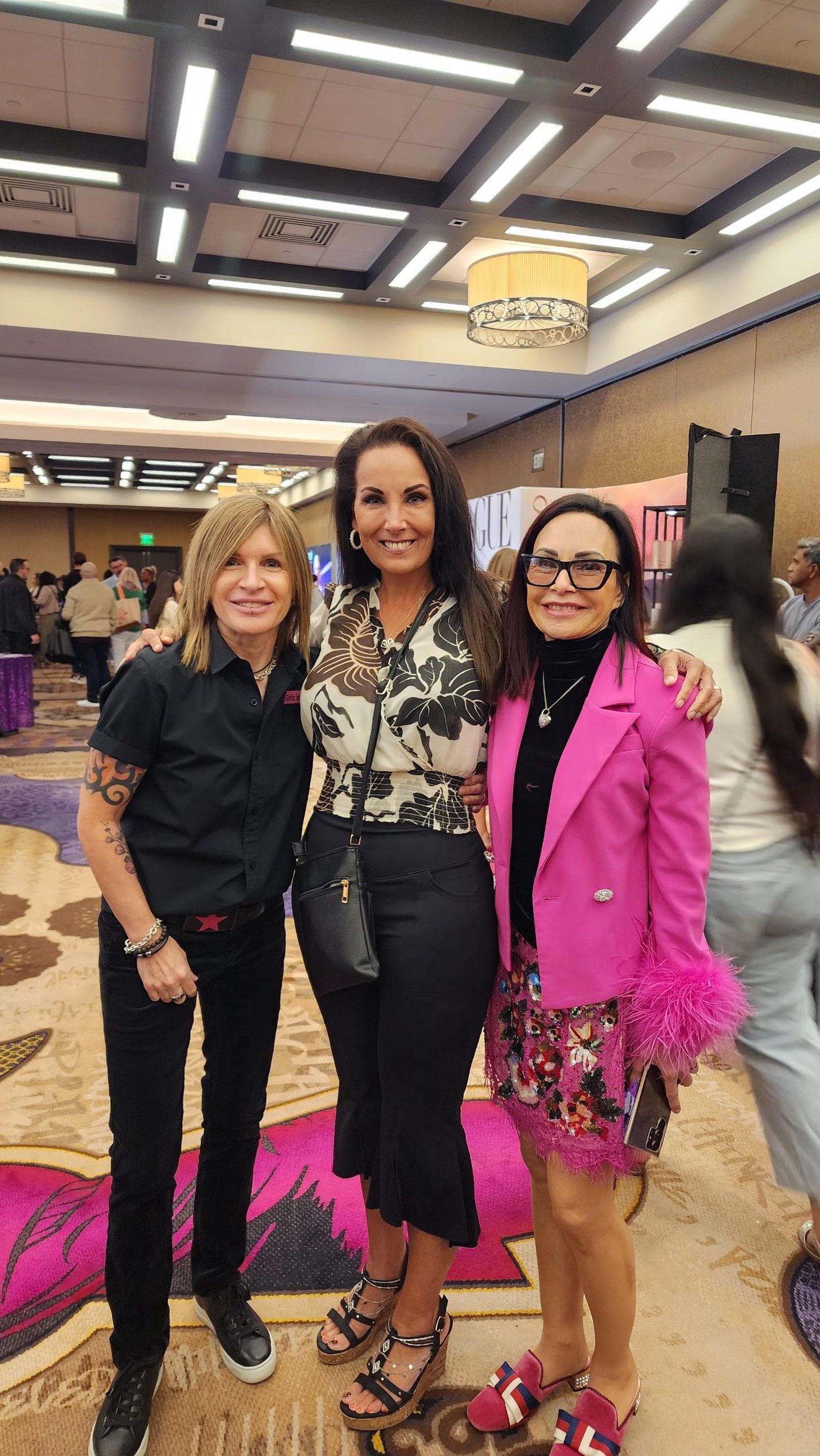 Three women pose together indoors. One wears a black shirt, another a floral top and black pants
