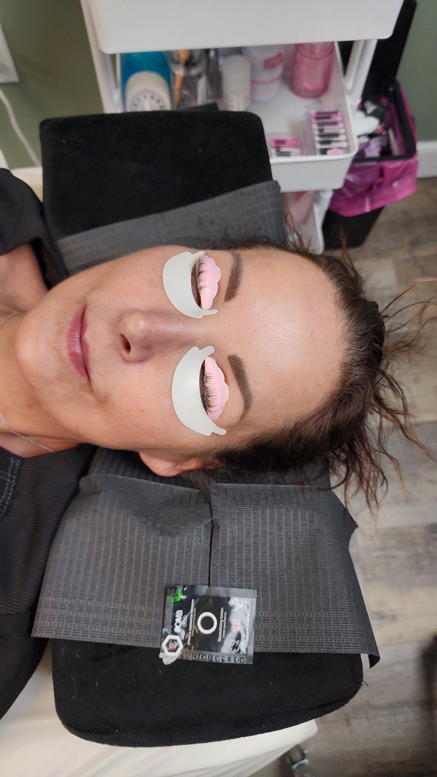 Woman with eye pads on, receiving lash extensions at a salon. She is lying down, smiling.
