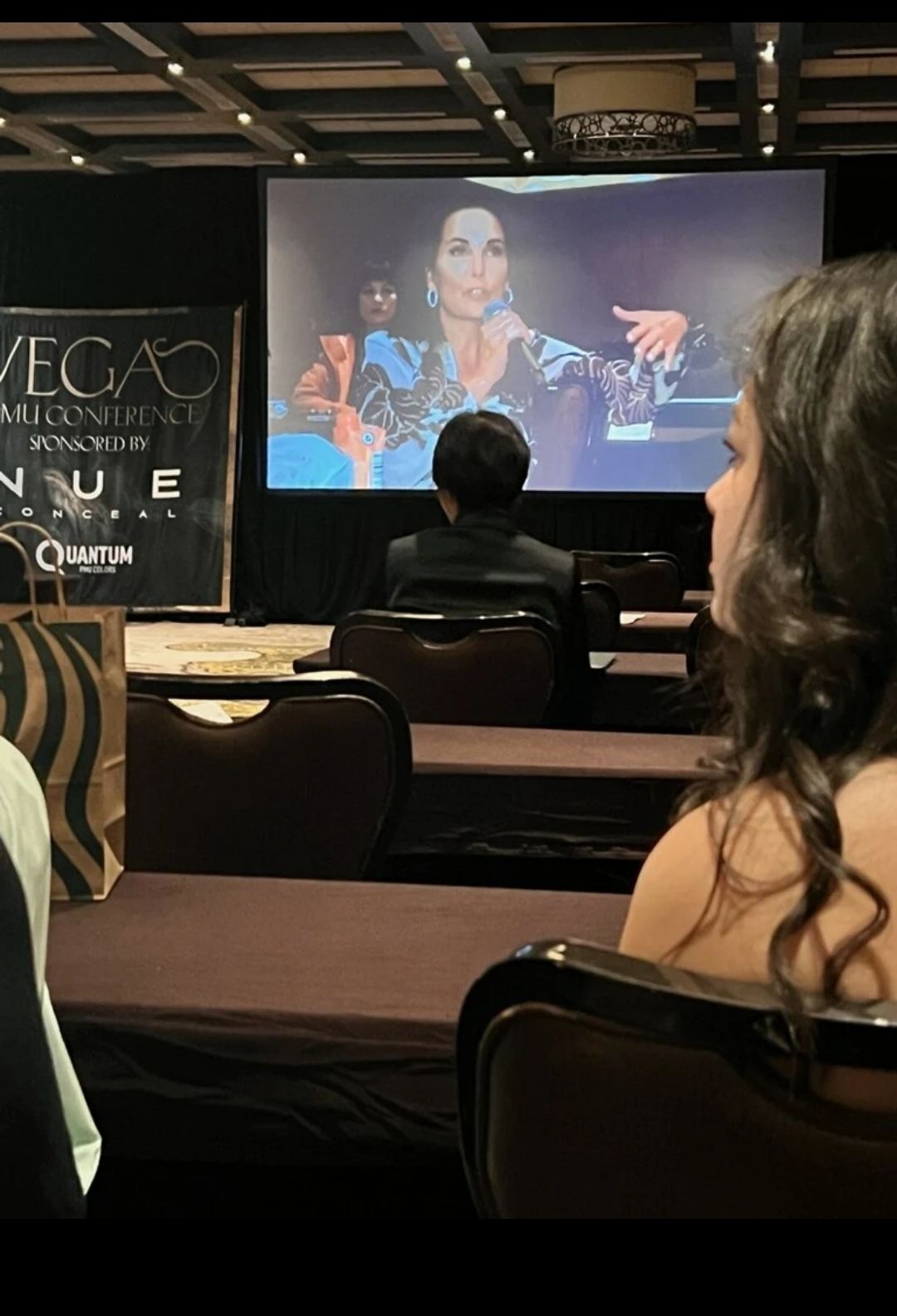 Audience watches a large screen displaying a woman speaking. Rows of chairs are arranged in a dimly lit conference room.