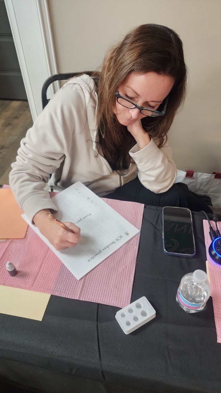 Woman with glasses writing on a paper, possibly designing a tattoo, with a phone and ink supplies on a table.