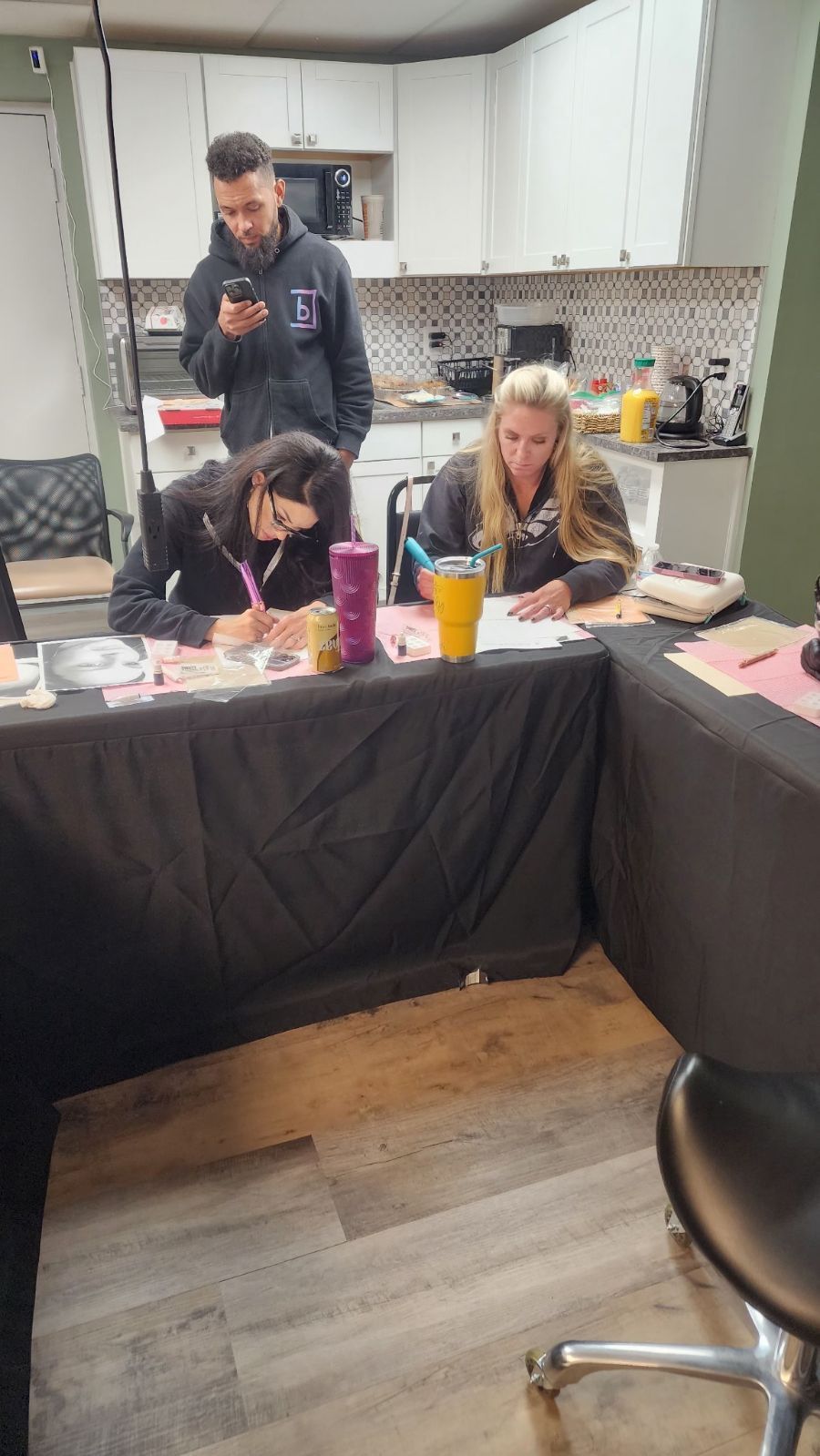 Three people at a table; two women are writing, a man is on his phone behind them in a kitchen.