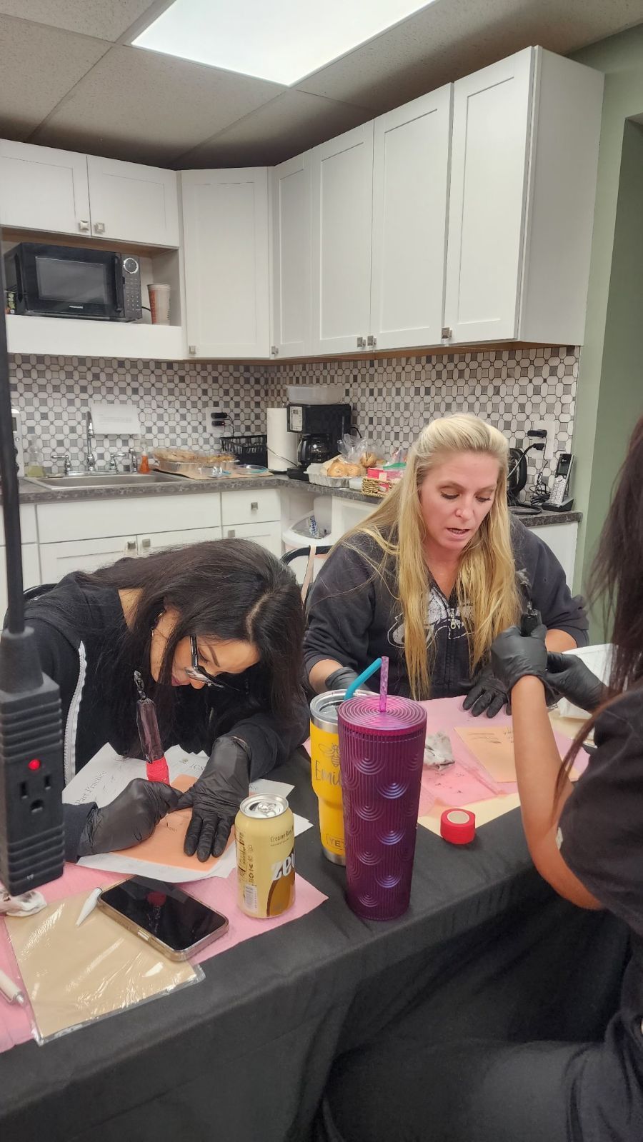 Two women wearing gloves practice tattooing on fake skin at a table.