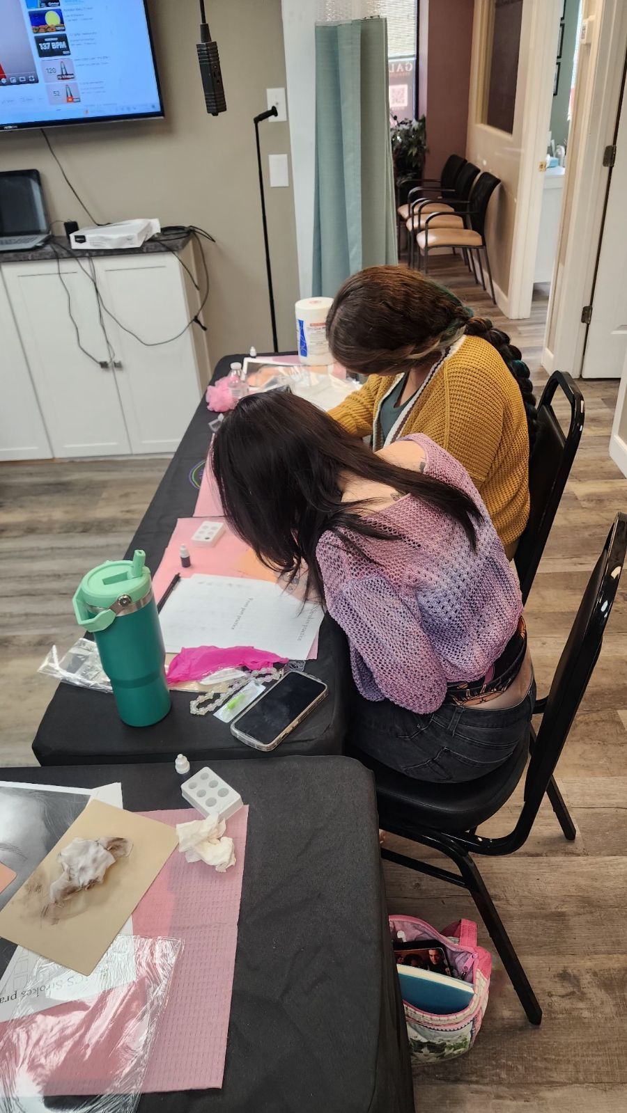 Two women seated at a table, working on projects. They are in a room with a TV, cabinets, and other supplies.