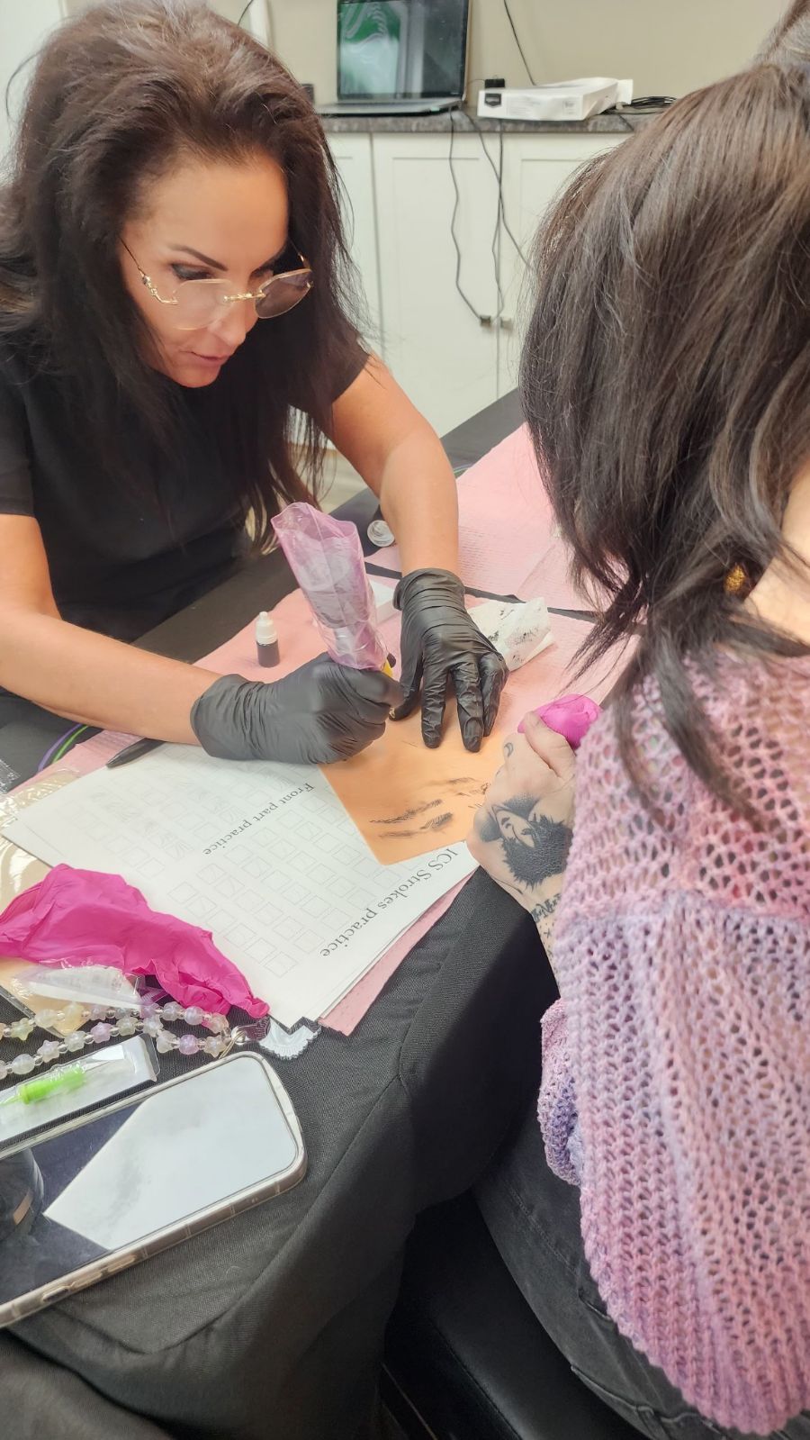 Woman in black gloves performing microblading on a client's eyebrow, indoors. Both are seated at a table with supplies.