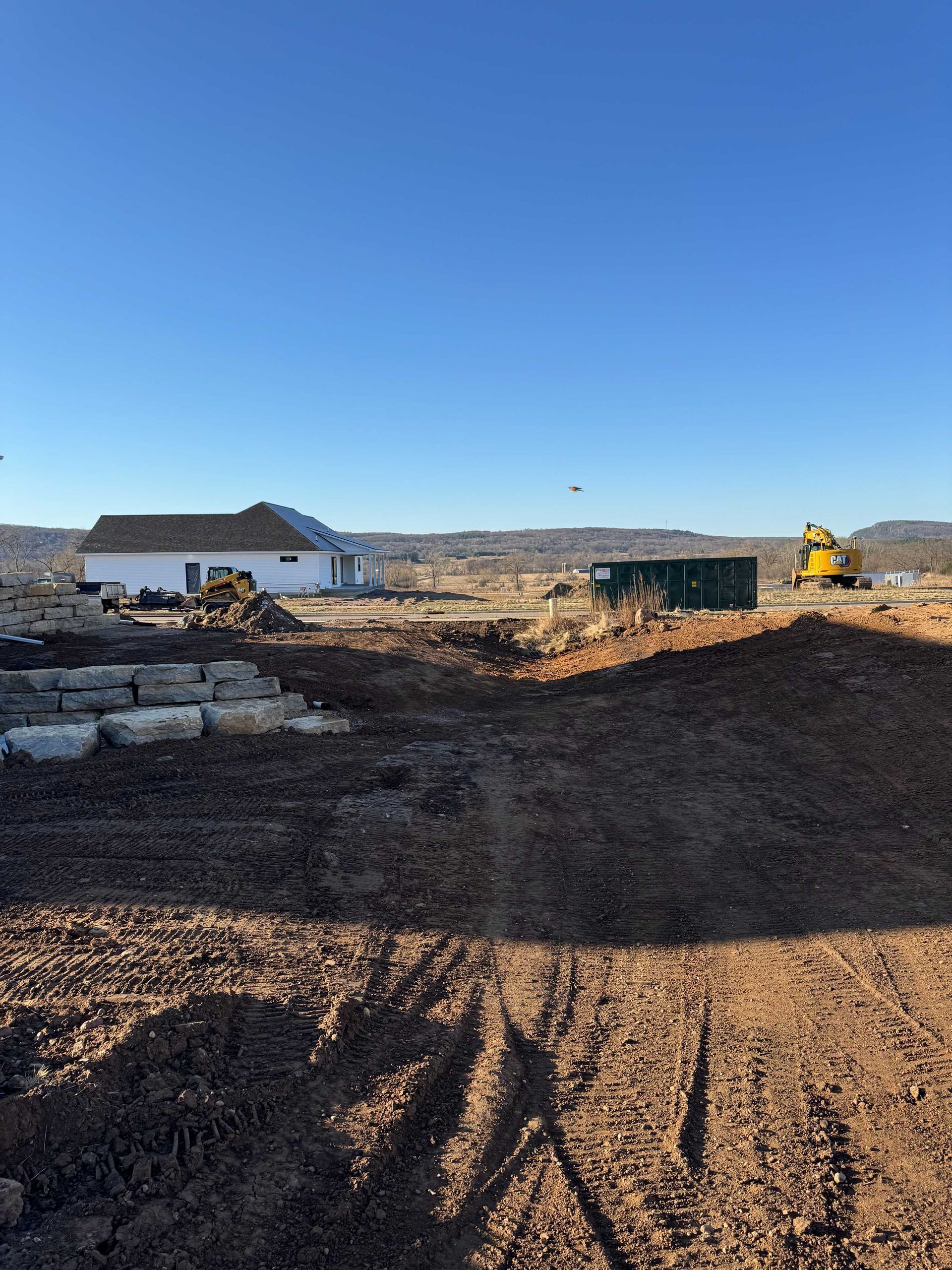 A construction site with a white house, large stone blocks, a green dumpster, and a yellow excavator under a blue sky.