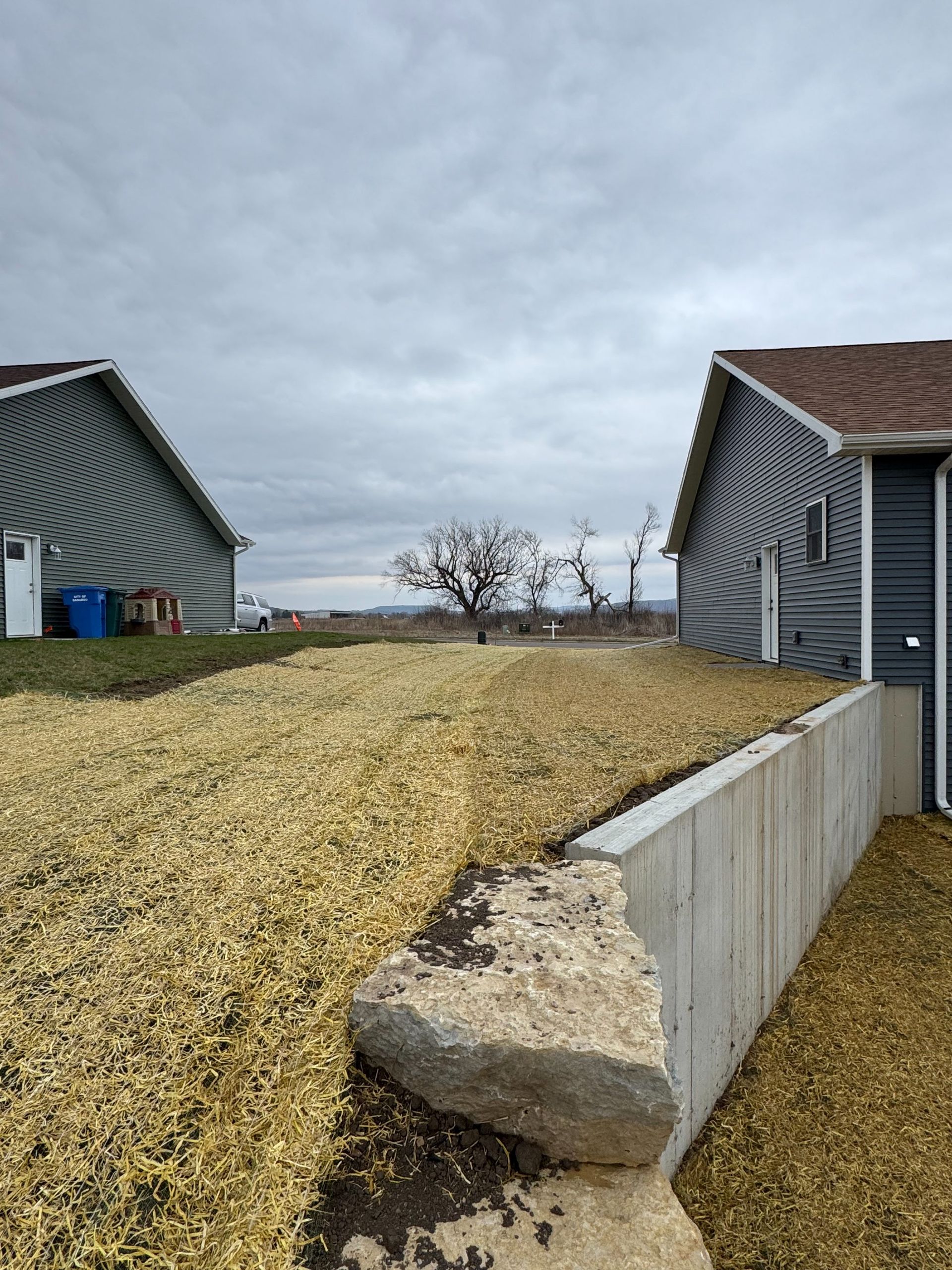 A concrete retaining wall separates two suburban homes with gray siding, with a gravel yard area between them.