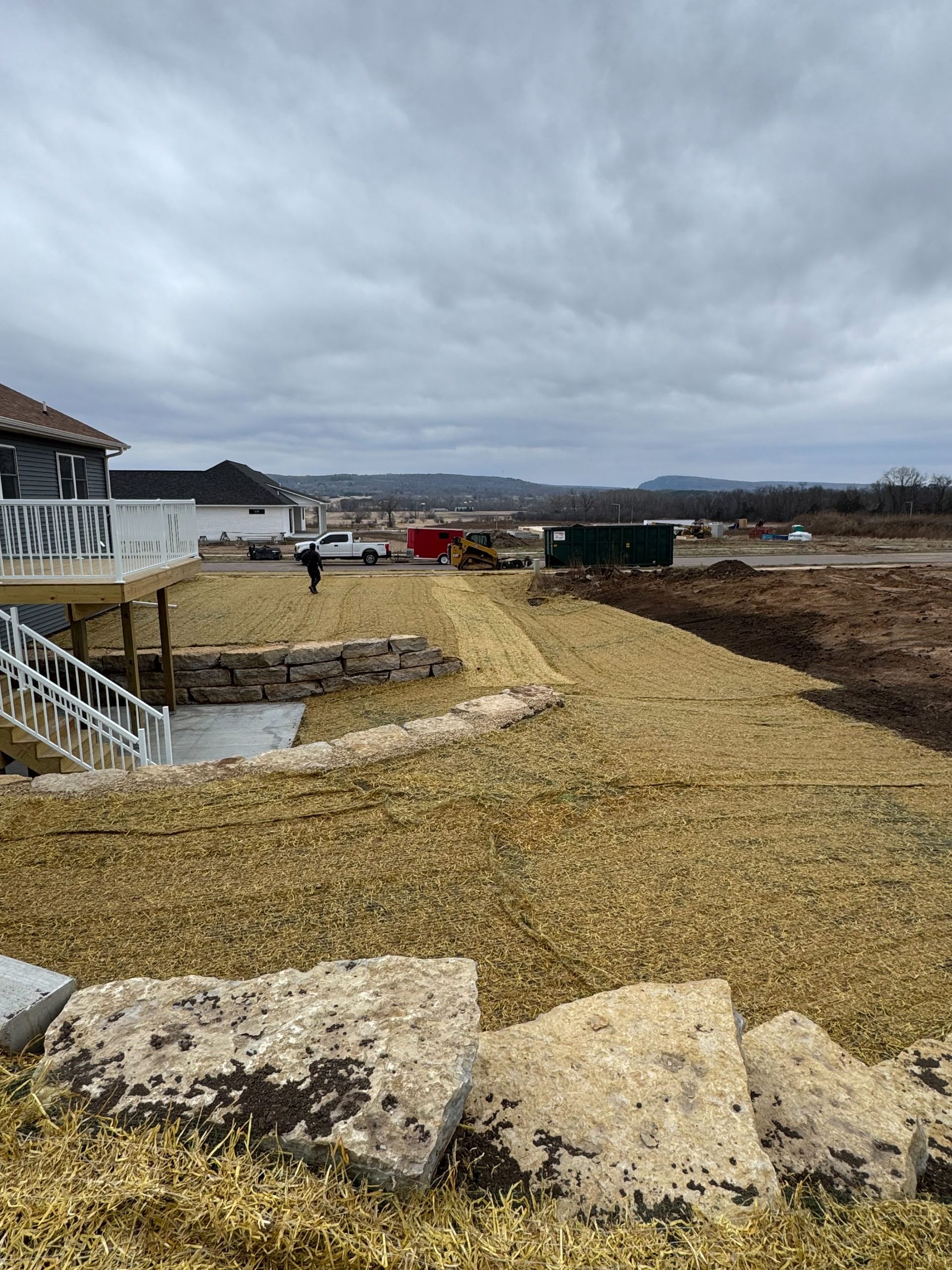 A residential backyard under construction with fresh straw mulch, stone retaining walls, a deck, and distant earthworks.