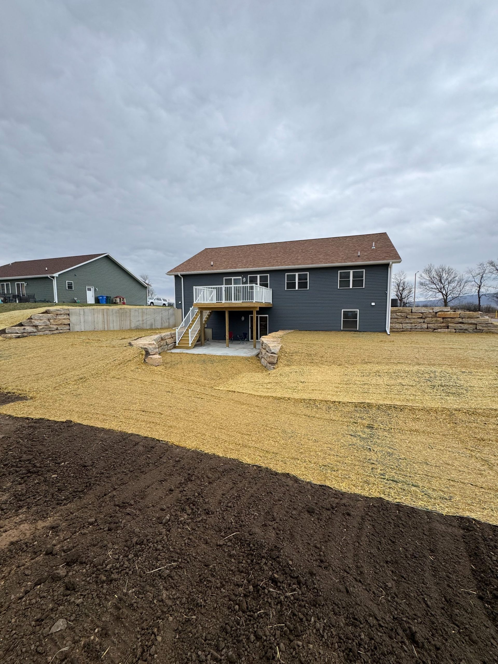 A backyard view showing a gray two-story house with a wooden deck, straw-covered ground, and a dark soil patch in front.