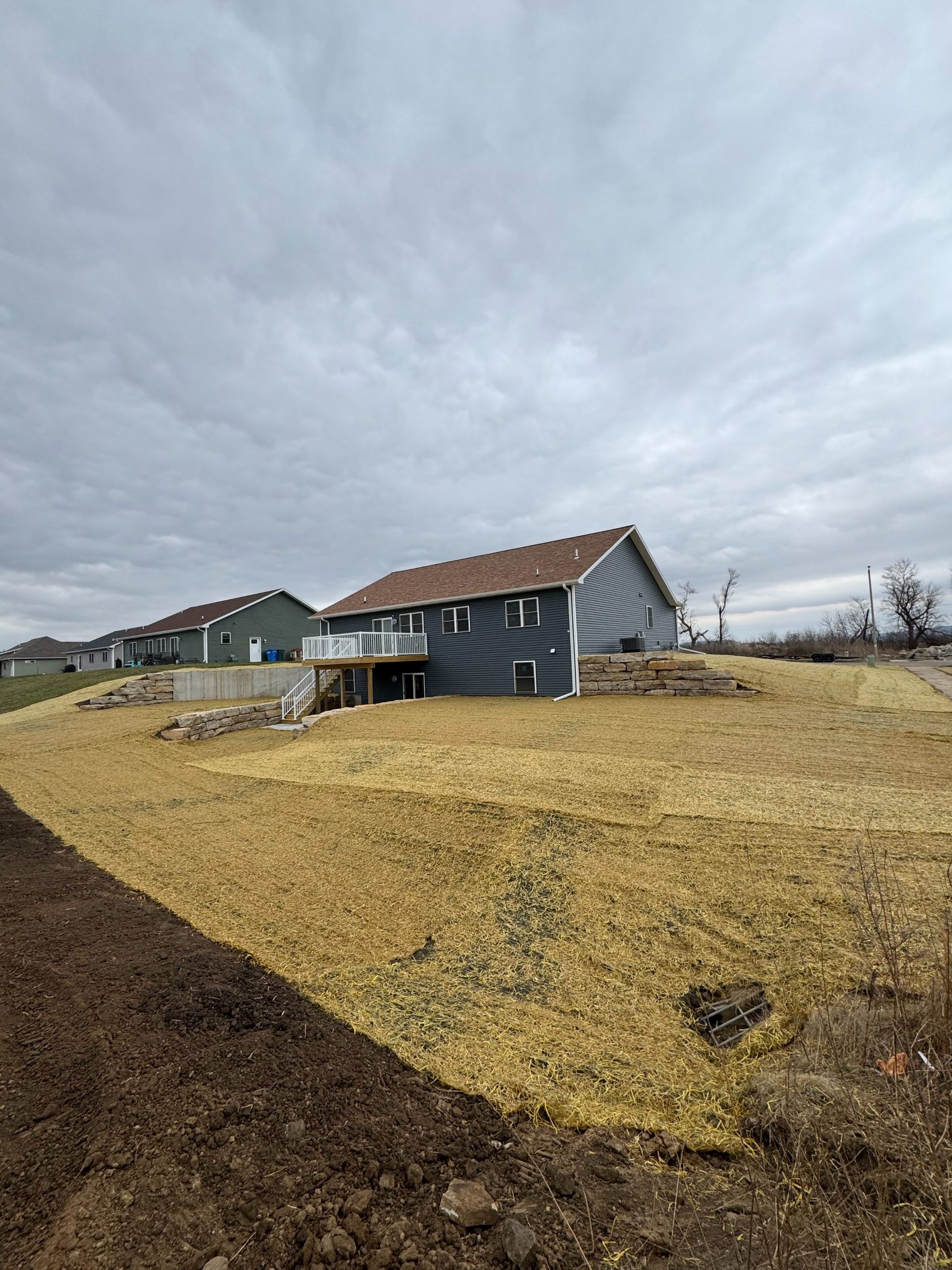A gray house with a wooden deck stands above a large, freshly mulched, and straw-covered terraced hillside.