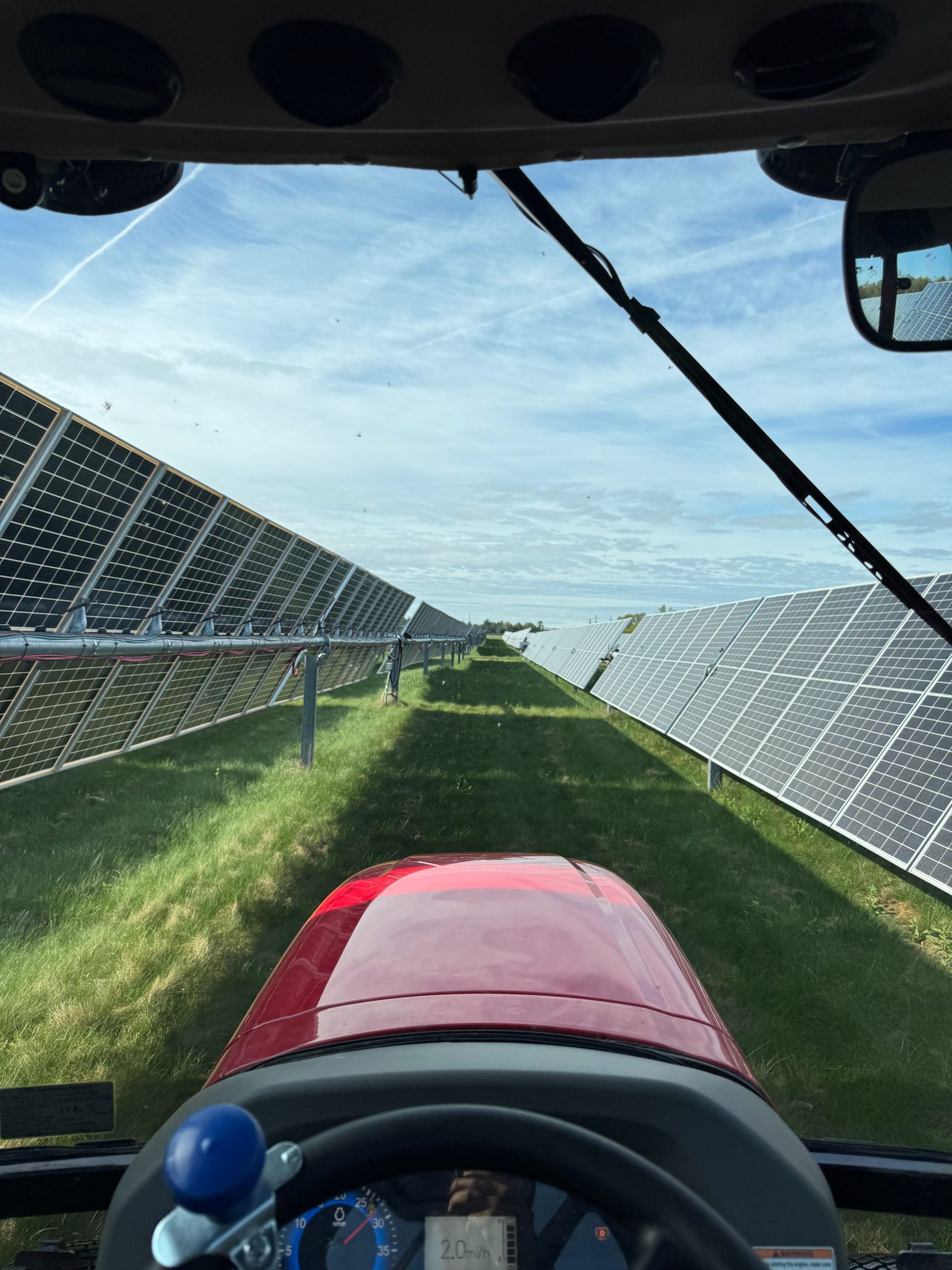 View from inside a tractor cab driving between rows of solar panels in a grassy field under a blue sky.